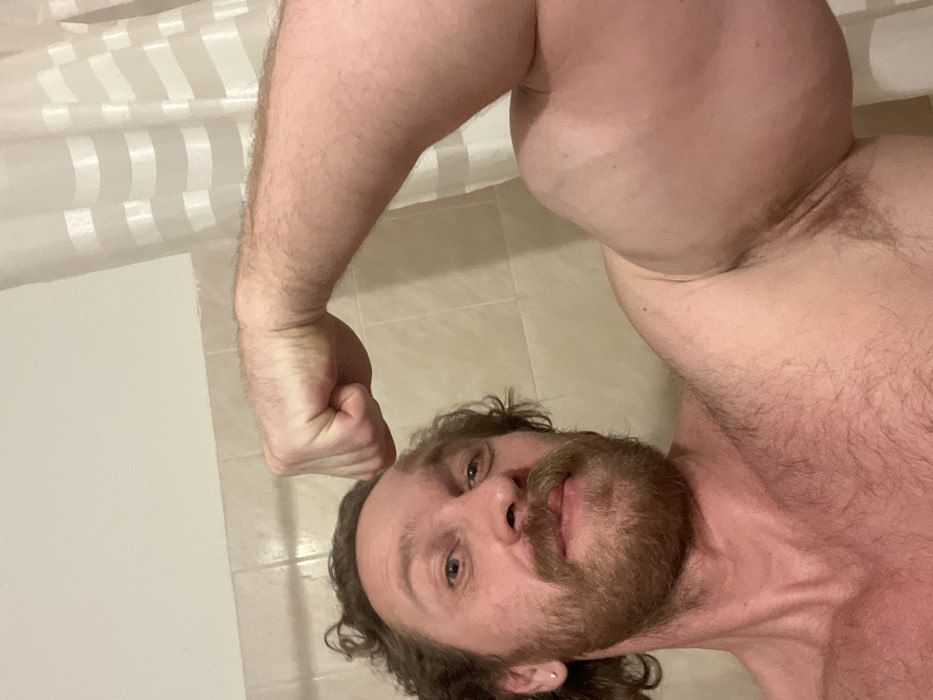 A shirtless man with a beard and mustache poses. He faces the camera with a slight smile. He's indoors in what appears to be a bathroom. His right arm is flexed, showcasing his bicep and forearm. A white shower curtain with vertical stripes is visible in the background. The bathroom has light-colored tiles on the wall and floor.