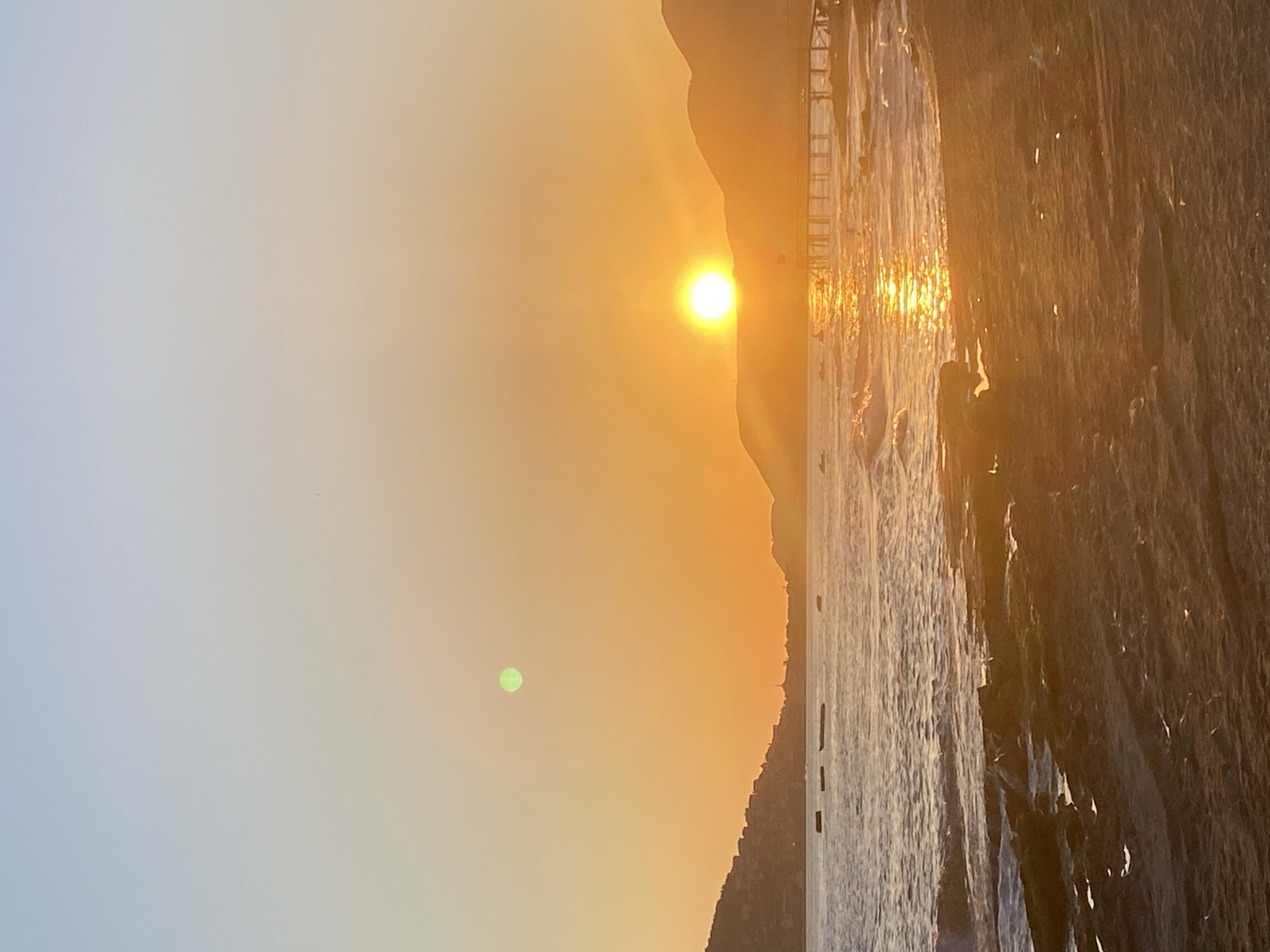 The image depicts a sunset over the Mediterranean Sea in Alanya, Turkey. The sun is positioned near the horizon, casting an orange glow over the sky and reflecting off the water. A mountain range is visible on the right side of the frame. A green lens flare is visible in the sky. The shoreline is rocky and rugged. A pier is visible on the right of the horizon.