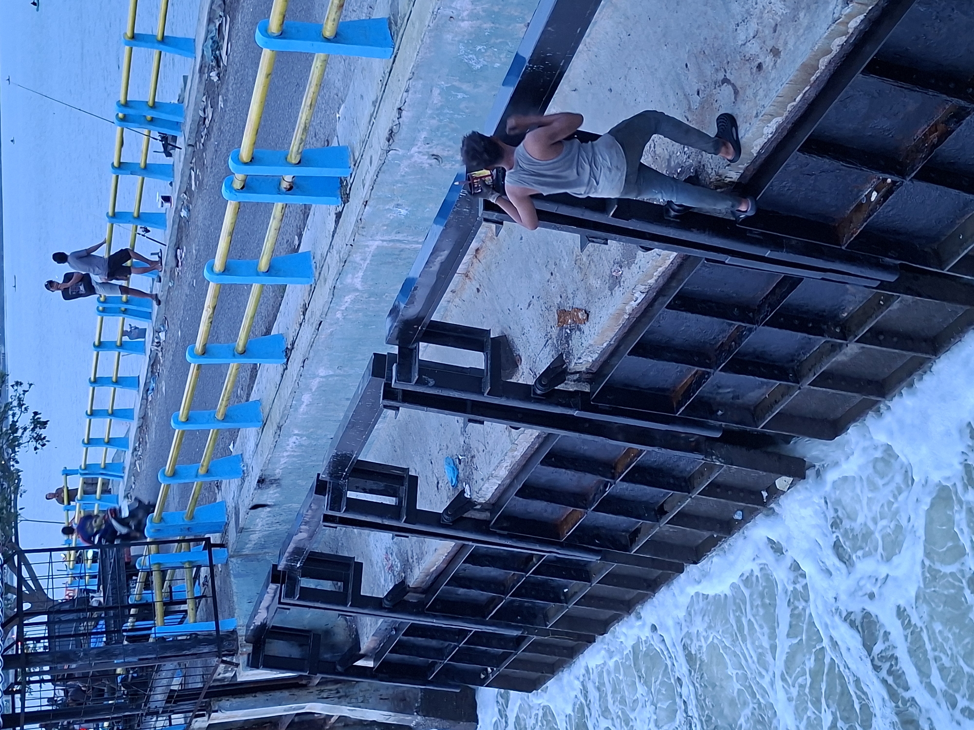 The image shows a waterscape scene, seemingly in Lhokseumawe, Indonesia, based on visual clues. A man is lying down, looking at a mobile phone, on what appears to be a structural part of a water management system, possibly a dam or floodgate. The structure is made of dark metal with a grid-like pattern above the water. Rushing water is visible beneath the structure.
In the background, there's a blue and yellow railing along a pedestrian area. Two men appear to be fishing off the railing with rods.