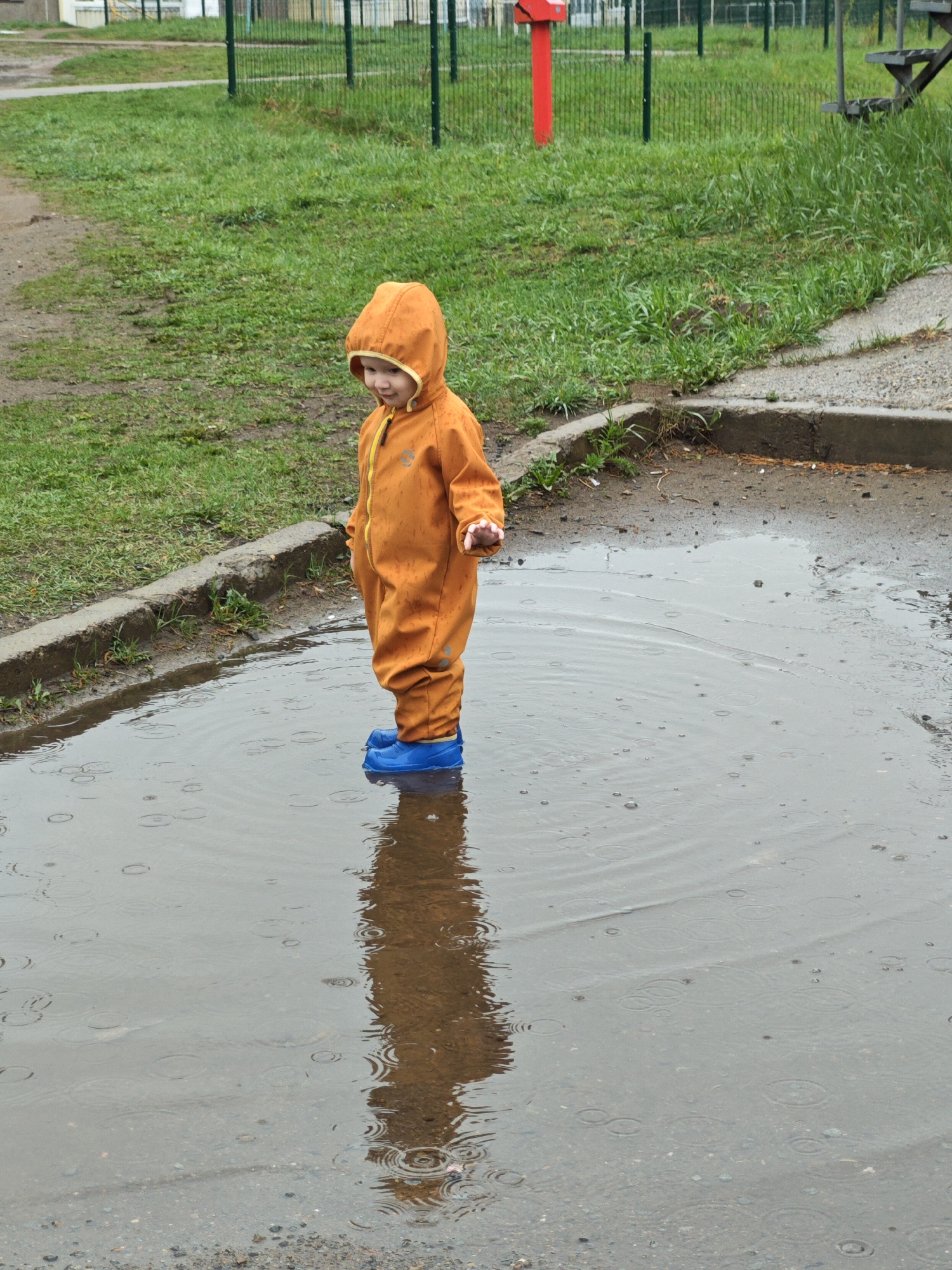 A small child in an orange hooded jumpsuit and blue boots stands in a large puddle. The child is positioned in the lower-middle of the frame, with their reflection visible in the water. The puddle takes up the foreground and extends into the middle ground. The ground around the puddle is wet, with some grass and concrete bordering it. In the background, a green fence and a red structure are visible. The location is possibly in Markovskii, Russia. It appears to be a rainy day.
