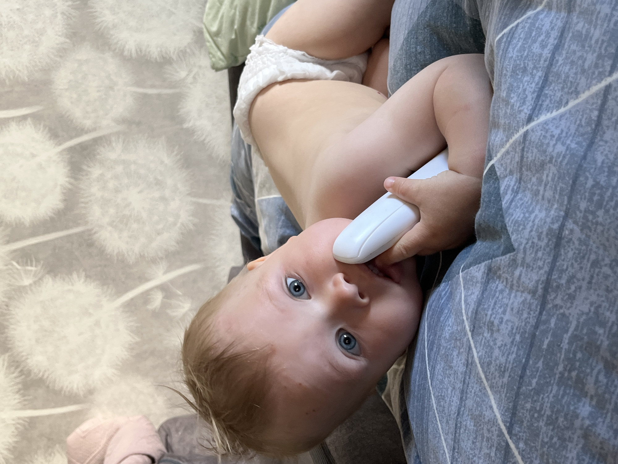 A baby is lying on a bed. The baby is wearing a diaper and holding a white object in its mouth. The baby has blue eyes and light brown hair. The bed has blue and grey striped bedding. The wall behind the baby has a white dandelion-patterned wallpaper. No information is provided in the picture to ascertain the location in the city of Chaikovsky, Russia.
