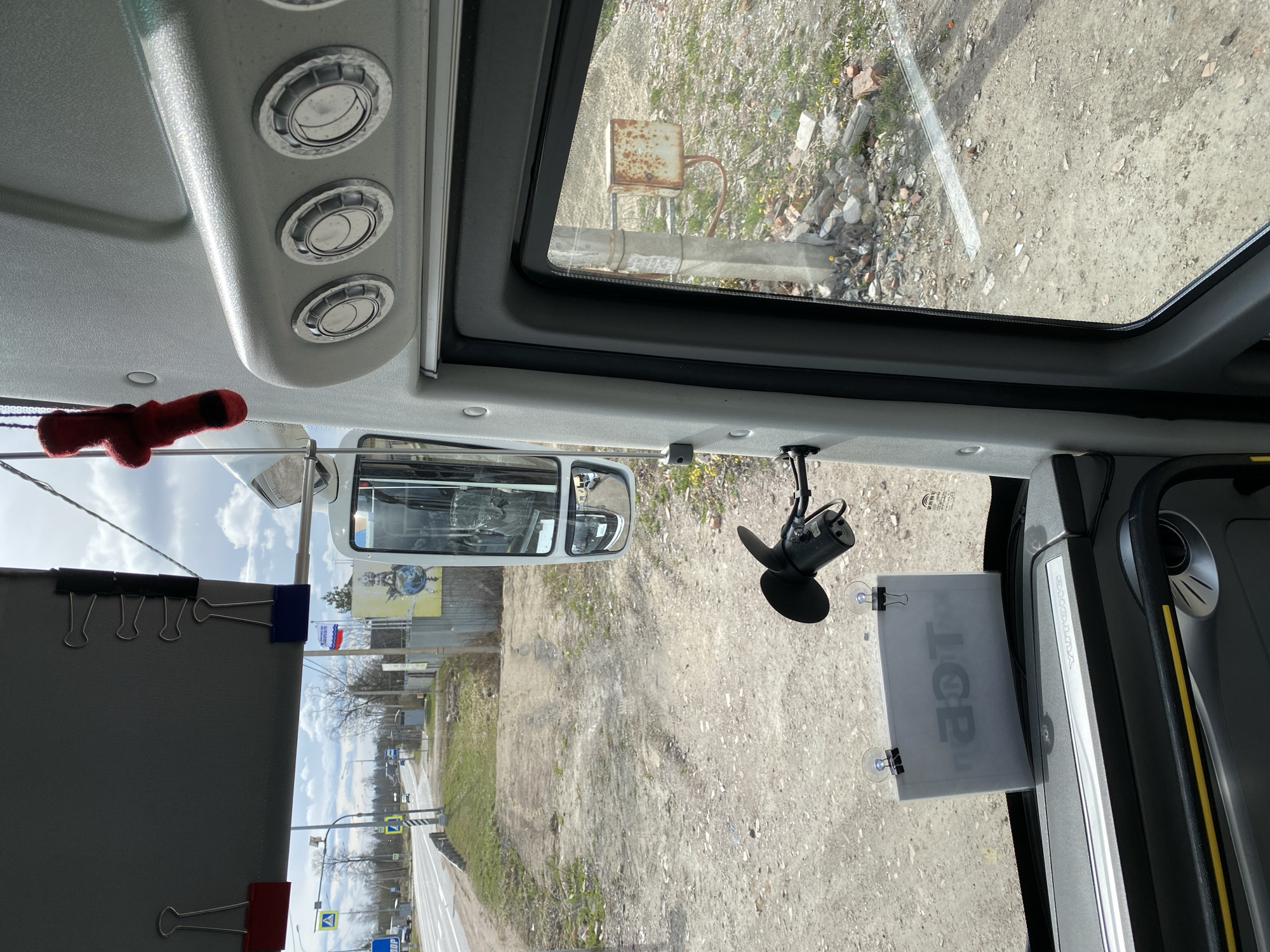 This image captures the interior view from a vehicle, likely a bus, looking out through the front and side windows. The perspective is from the driver's area, facing forward and slightly to the right.
Inside the vehicle, three circular air vents with metallic finishes are visible on the ceiling panel above. A small red, plush object, possibly a toy or decoration, hangs from the ceiling near the rearview mirror. The rearview mirror itself reflects part of the vehicle's interior, showing seats or interior structures. A black device, resembling a microphone or sensor, is attached to the windshield near the top right. A translucent sign or document is affixed to the passenger side of the windshield with suction cups and black binder clips, displaying partially visible Cyrillic text, "ПОР" (likely part of a word like "ПОРЯДОК" meaning "order" or "procedure"). Another grey fabric panel, possibly a sunshade or curtain, is held by binder clips on the left side of the windshield frame. On the vehicle's dashboard or side panel, the brand name "YUTONG" is visible.
Outside the vehicle, the scene reveals an unpaved, dusty ground area with scattered debris like rocks and broken concrete, along with patches of sparse grass. In the distance through the top window, a rusty metal box is mounted on a pole near what appears to be a concrete barrier. Through the main windshield, a road with a crosswalk sign and a main road sign (blue square with a yellow arrow) is visible further away. Utility poles and wires crisscross the sky. Several small flags, one clearly the Russian flag and another blue and white, are visible hanging from a structure in the mid-ground, alongside a banner depicting the Earth from space on a yellow background. The sky is bright and clear, indicating daytime and good weather.
The scene suggests the vehicle is either stationary or moving slowly through a semi-rural or industrial area in Bol'shoe Teshkovo, Russia.