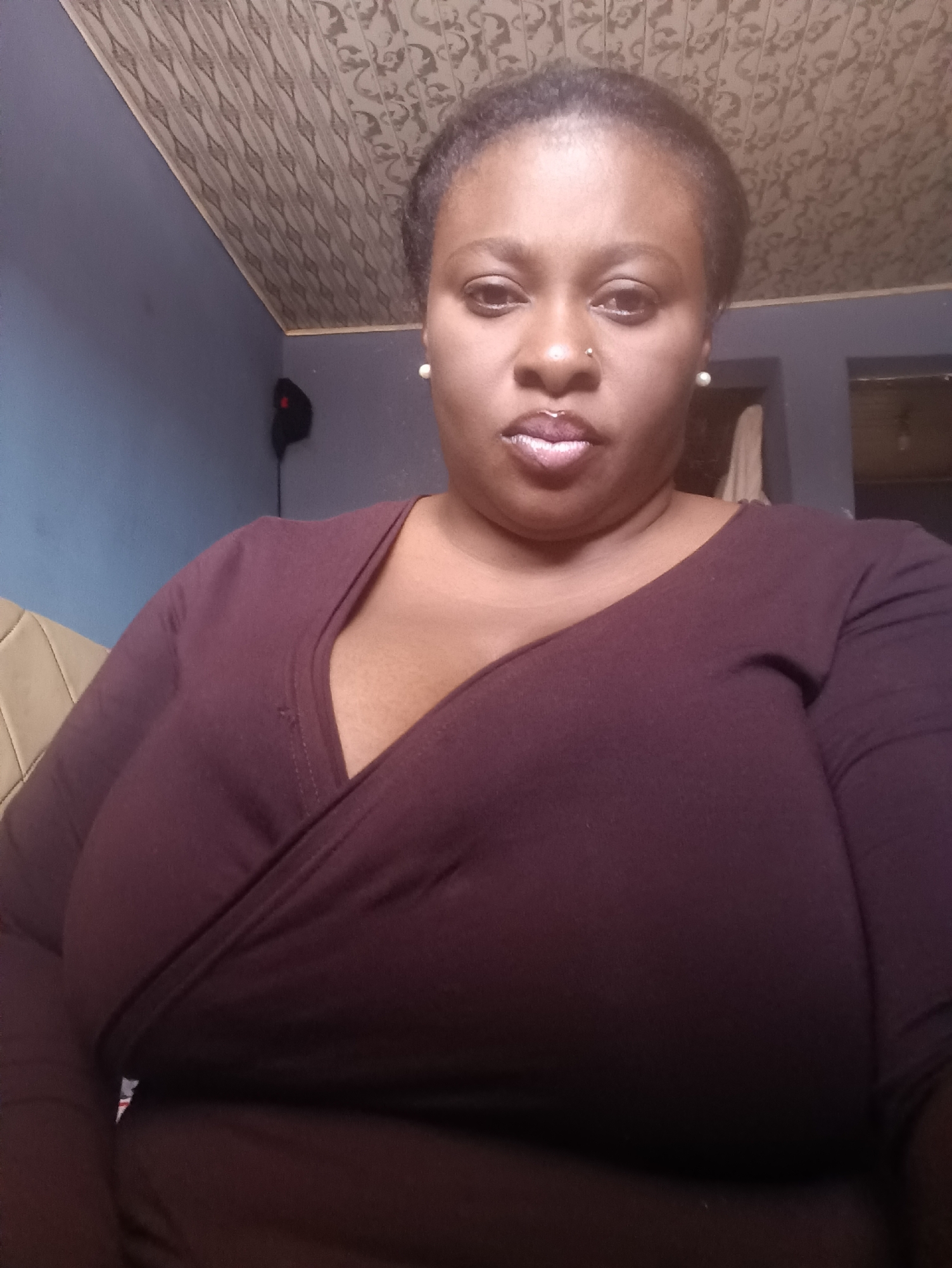 A medium shot depicts a woman indoors. She has dark skin, dark hair styled back, and is wearing pearl earrings and a nose ring. Her lips appear glossy. She is wearing a long-sleeved, maroon-colored wrap-style shirt. The background includes a blue wall, a patterned ceiling, a section of a beige couch on the left, and a doorway or window on the right. A black object is attached to the wall behind her on the left. Location is unknown, and no information suggests it is in Jos, Nigeria.
