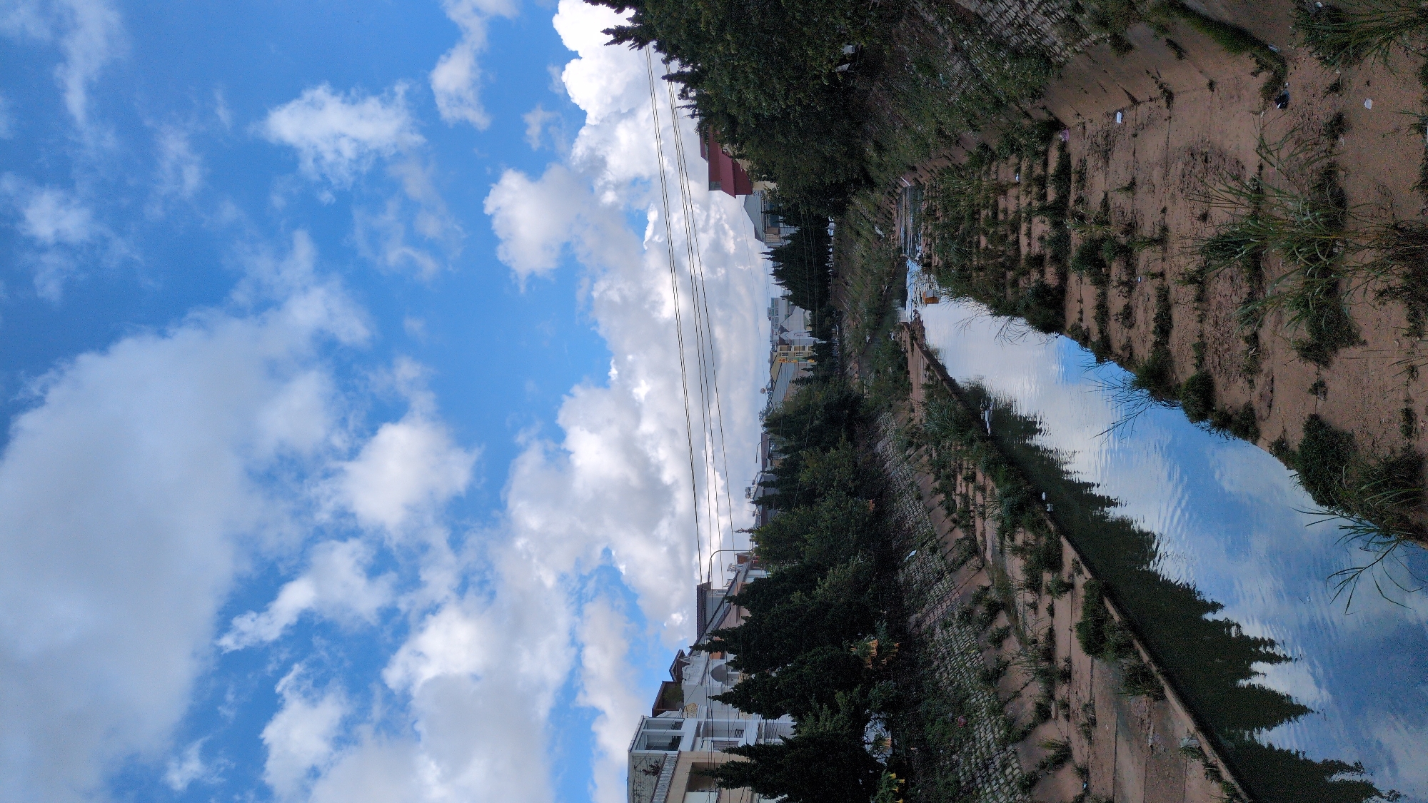 The image shows a canal running through a residential area in Da Lat, Vietnam. The canal is concrete-lined, with grassy banks and some scattered debris. Trees line the sides of the canal, and houses and buildings can be seen in the background. The sky is blue with white clouds. The canal appears to be a drainage channel or a man-made waterway, possibly for flood control or water management within the urban landscape. The presence of residential buildings suggests this is an inhabited part of the city.