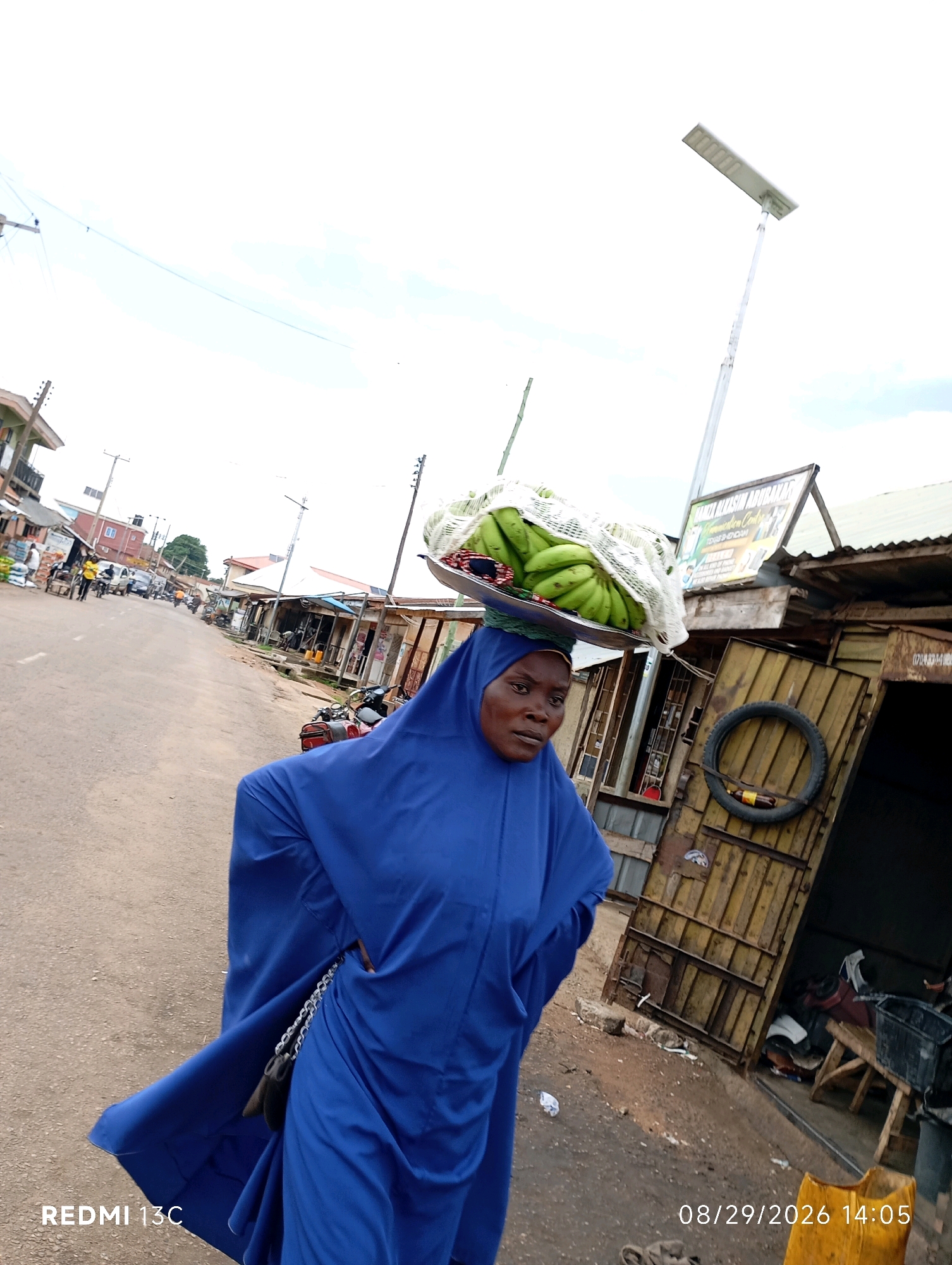 In Shendam, Nigeria, a woman wearing a blue hijab and a flowing blue garment walks on a paved road. She carries a tray of green bananas balanced on her head, partially covered by a white mesh bag. The street is lined with buildings, some appearing to be shops with corrugated metal roofs. A solar-powered street lamp is visible in the background. The scene suggests a busy marketplace or commercial area, with other pedestrians and motorbikes present on the road.