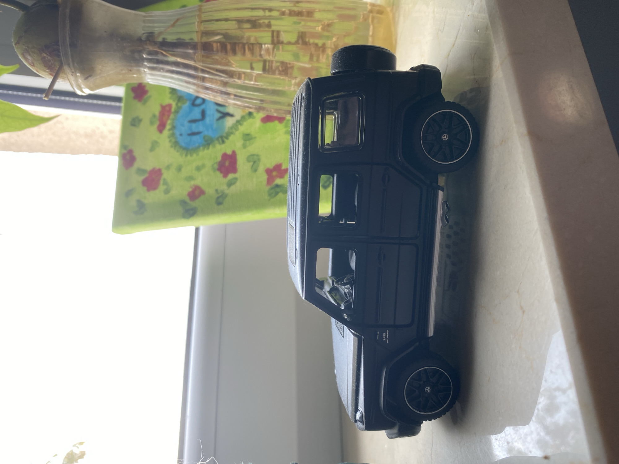 A black toy Mercedes-Benz G-Wagon is parked on a white, reflective surface, possibly a windowsill. The toy car is detailed, featuring visible wheels, tinted windows, and a spare tire mounted on the back. Behind the car, a green canvas painting with a heart shape and floral designs is partially visible. To the right, a clear glass vase with a plant and water is also present. The background is out of focus, showing what appears to be a window with bright light filtering through. The scene is indoors, with a distinct shadow cast by the toy car on the surface below. There are no people or animals present. The location is not specified as Łódź, Poland.