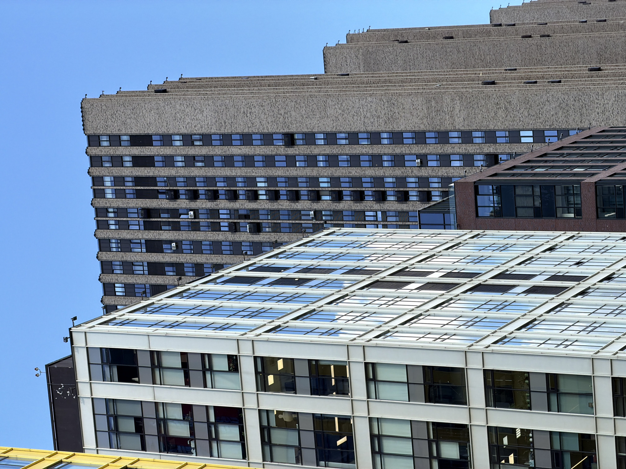 This image captures a complex architectural scene in Khimki, Russia, featuring a composition of multiple modern and brutalist-style buildings under a clear blue sky.
In the immediate foreground, at the bottom of the frame, a glimpse of a building with a bright yellow roof is visible. Above this, a prominent white building dominates the lower-middle section. It features large, rectangular windows with light-colored frames, some revealing interior office spaces with lights on, while others reflect the sky. The roof of this building is a striking glass structure, composed of many rectangular panes arranged in a grid pattern, reflecting the blue sky and adding a bright, geometric element to the scene.
Behind and to the right of the white building, a brown brick building with large, dark-framed windows is partially visible. This building also incorporates a glass roof or skylight section, similar in structure but darker in tone than the white building's roof.
The most dominant structure in the background is a massive, multi-tiered building with a rough, textured, grey-brown concrete or stone facade, indicative of a brutalist architectural style. This building features numerous small, rectangular windows arranged in horizontal bands across its many floors. Its upper sections recede in a distinctive stepped pattern, with the tiers becoming narrower towards the top. Several small, dark objects are sparsely scattered along the edges of these upper tiers. Many of the windows on this building appear dark, though some reflect light or show a lighter interior.
The overall impression is one of urban density and architectural contrast, with the clean lines and reflective surfaces of the modern glass buildings juxtaposed against the heavy, textured mass of the brutalist structure. The bright sunlight and clear blue sky suggest a daytime setting with fair weather. No people or visible text are present in the image.