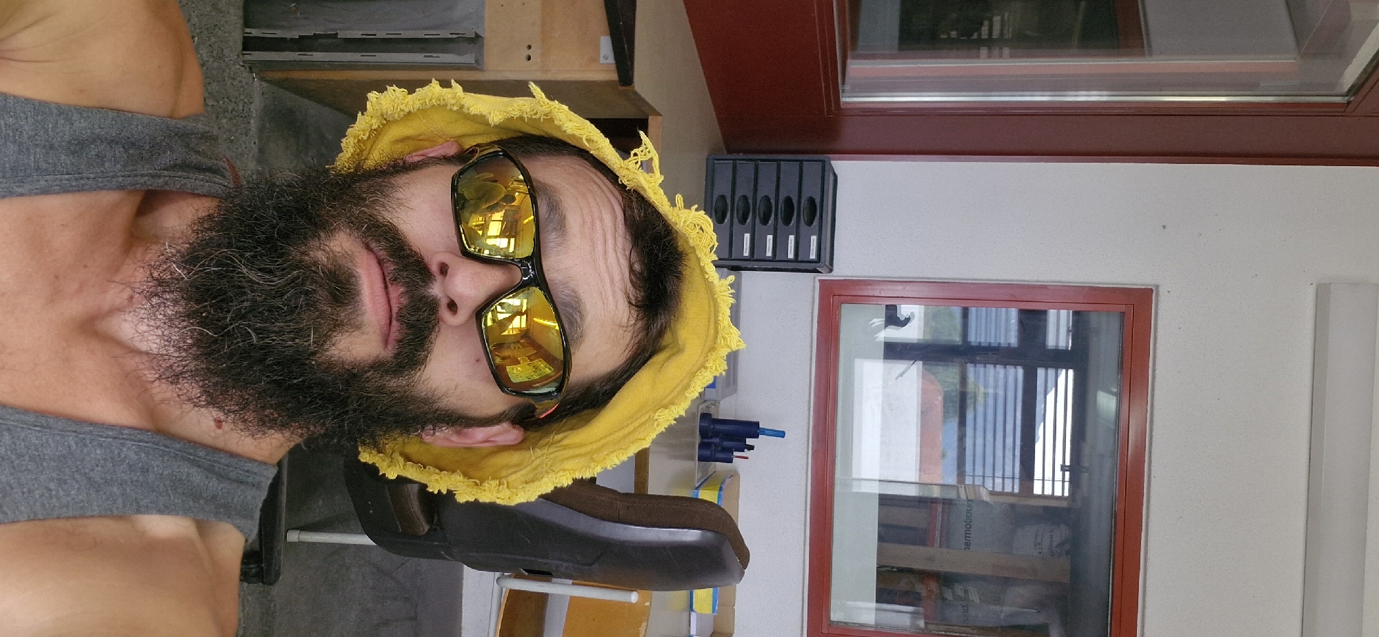 A close-up, eye-level, selfie-style photograph features a man with a full beard and a yellow bucket hat with frayed edges. He is wearing reflective sunglasses with orange accents and a grey tank top. The background appears to be an indoor workspace or office, likely in Uetendorf, Switzerland, given the context provided. Elements visible include a large window with red framing, a filing cabinet, a desk, a rolling chair, and office supplies. The lighting suggests an indoor setting with natural light contributing to the overall illumination.