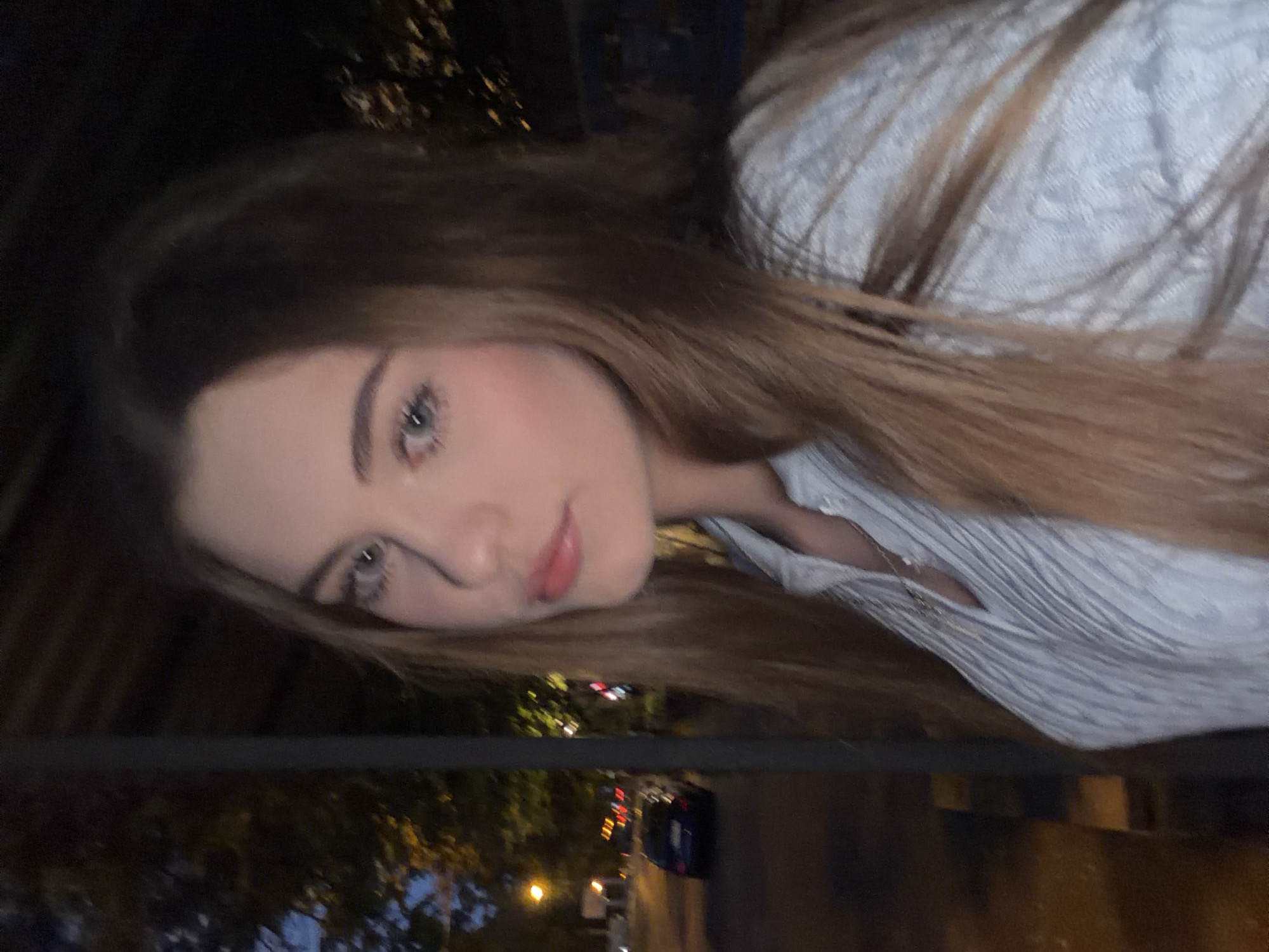 The image shows a medium shot of a young woman with long, light brown hair, looking to her left. She is wearing a light blue, collared shirt with a textured pattern and a thin necklace. Her makeup appears to include blush and lip gloss. The background is out of focus, showing an urban nighttime scene. To the left, a street with a dark blue car and streetlights is visible. Trees with illuminated foliage suggest a park or treelined street. The overall impression is that the photo was taken outdoors at dusk or after dark in a city. However, there are no specific visual cues to confirm the location as Dnipro, Ukraine.