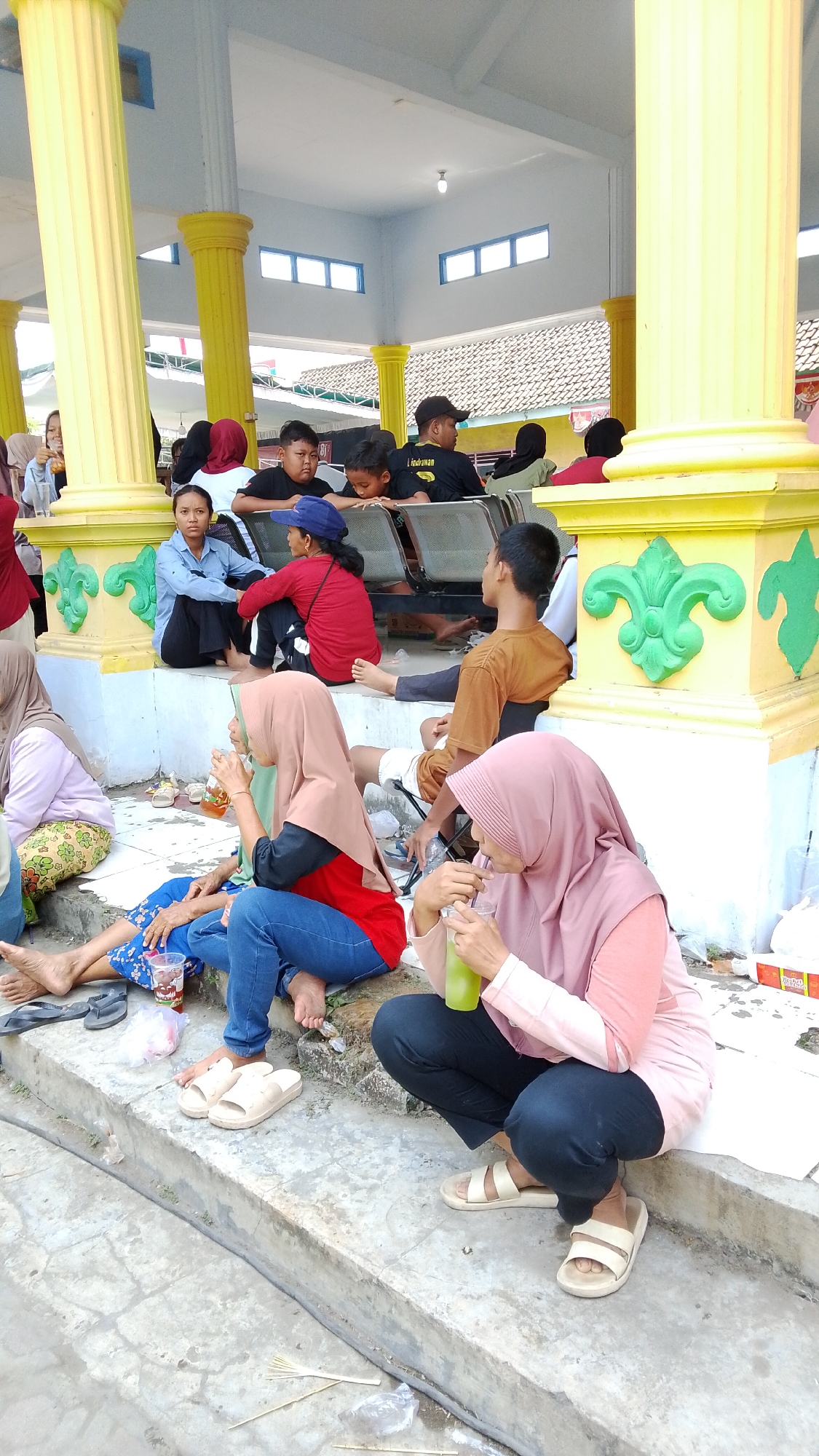 In Mancar, Indonesia, a group of people are gathered under a covered outdoor structure with yellow pillars and a white ceiling. Several individuals are seated on the ground or on steps, some of them women wearing hijabs. One woman in a pink hijab is drinking from a green beverage in a clear plastic cup with a straw. Another woman in a beige hijab is also holding a beverage. A child in blue pants and a floral top is lying on the ground. Other individuals, including children and adults, are visible in the background, some sitting on benches. The ground is paved with concrete, and there are scattered items like plastic bags and flip-flops. The overall atmosphere appears to be a casual gathering.