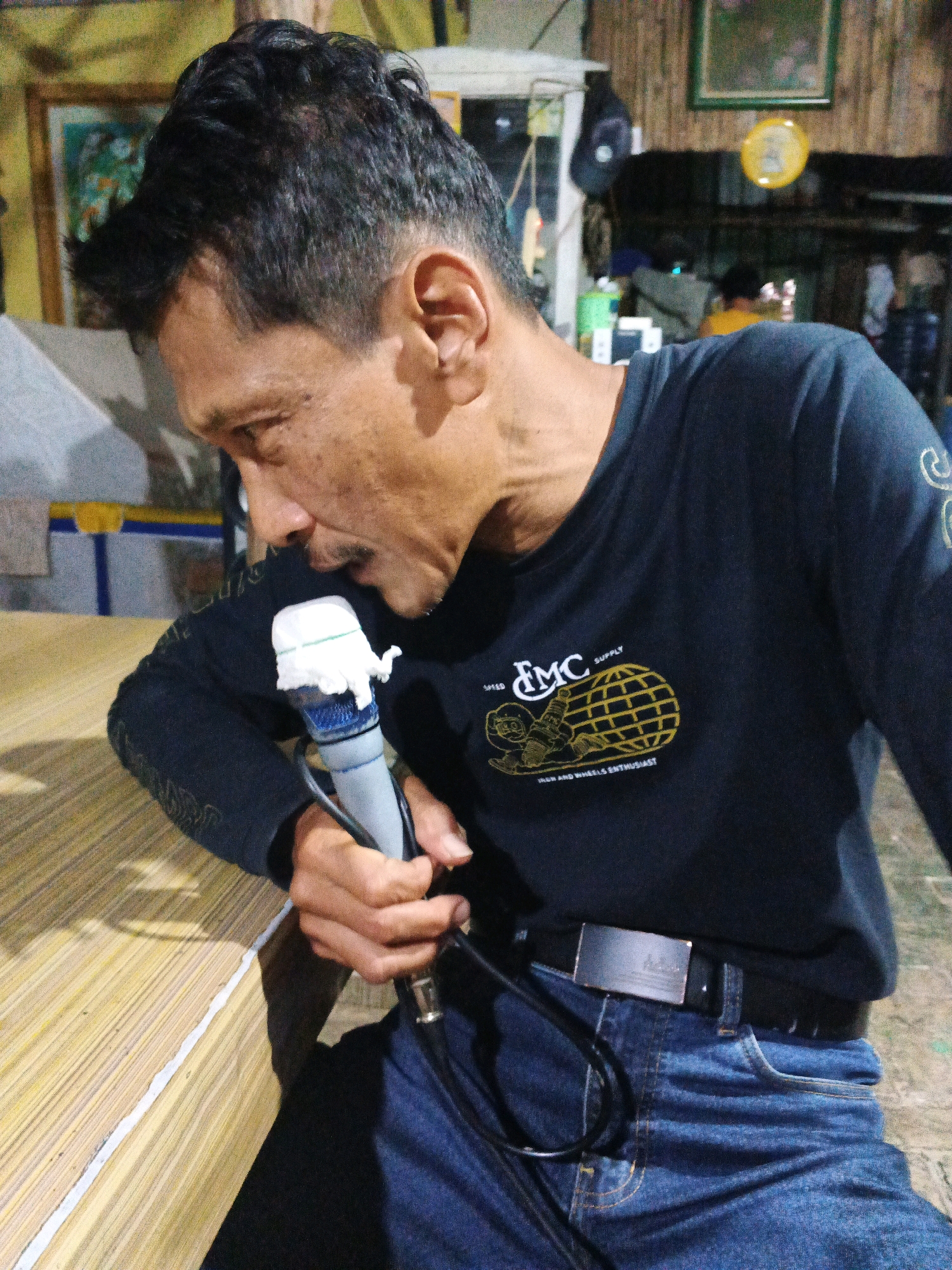 A medium shot captures a man speaking into a microphone in what appears to be an indoor, dimly lit setting in Ungaran, Indonesia. The man, facing right, is leaning forward slightly, his mouth close to the microphone. He has dark hair, styled with a fade, and a mustache. He is wearing a black long-sleeved shirt with a yellow logo on the left chest, and blue jeans with a dark belt. The microphone, held in his right hand, has a white covering on top, possibly a windsock or a makeshift cover. A cable extends from the microphone towards the foreground. The background suggests a casual environment, with hints of furniture, shelves, and framed pictures on a wall. The man is seated at a table made of woven material.