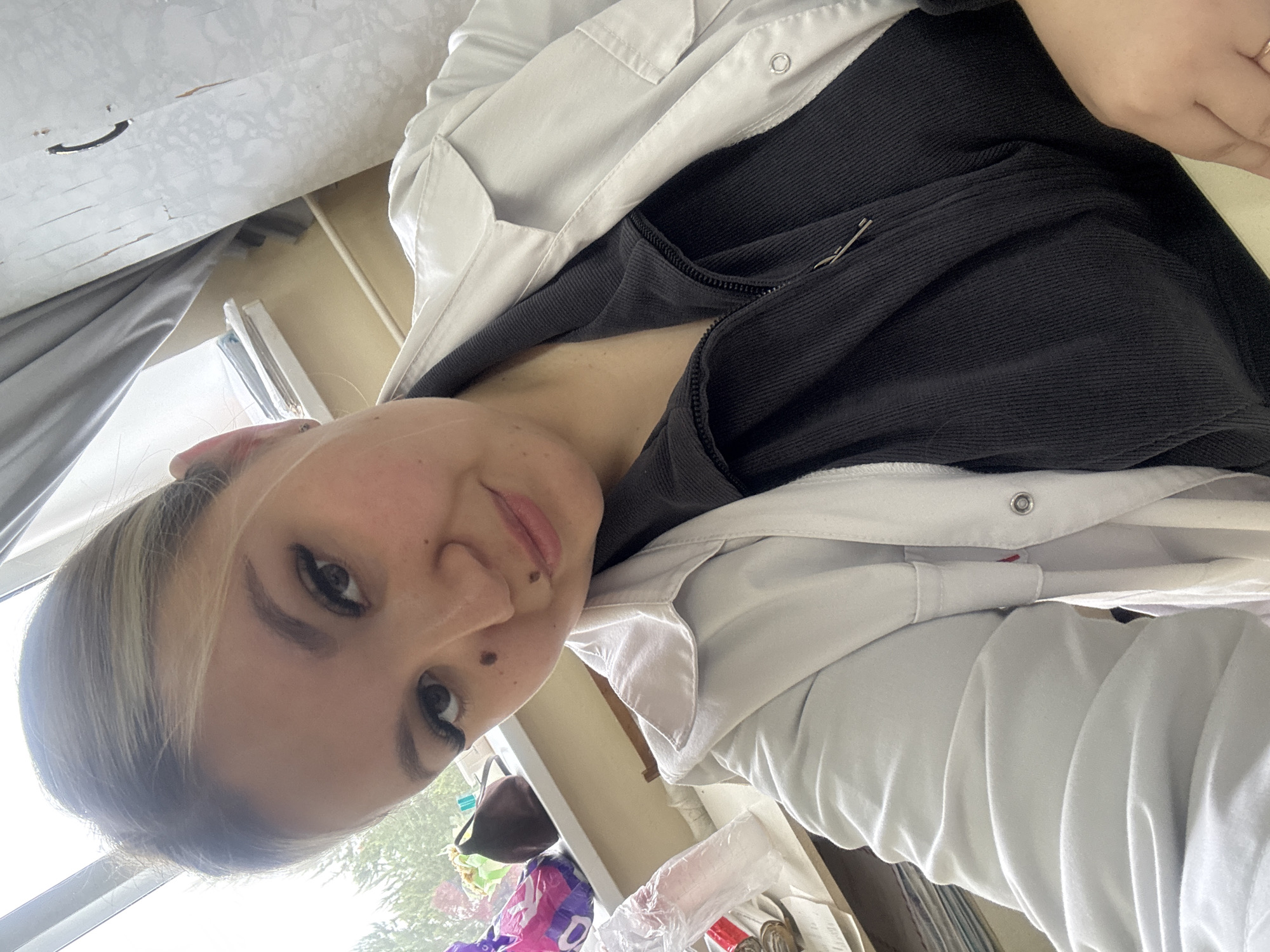 A young woman is depicted in a selfie-style photograph, taken at an upward angle, likely indoors during the daytime. Her light brown hair, with subtle blonde highlights, is pulled back. She has dark eyebrows and eyelashes, and several moles on her face, notably one below her left eye and another near her mouth. Her lips are a soft pink, and she wears a calm expression with a gentle smile.
She is dressed in a black ribbed top, possibly with a zipper, worn under a light-colored (white or light grey) jacket or lab coat. The jacket features pockets and a silver snap button on the lapel. A safety pin is attached to her black top near her left shoulder. A ring is visible on the ring finger of her right hand, which rests on her chest area.
The background shows a light-colored ceiling or upper wall with some dark marks or scratches. A grey curtain hangs to the left. Through a window behind her, bright natural light illuminates green foliage, indicating an outdoor view. On a surface beneath the window, various items are present, including clear plastic containers or cups and a partially visible purple plastic bag with an indiscernible white pattern. The setting appears to be a casual indoor environment. No clearly legible text is visible in the image.