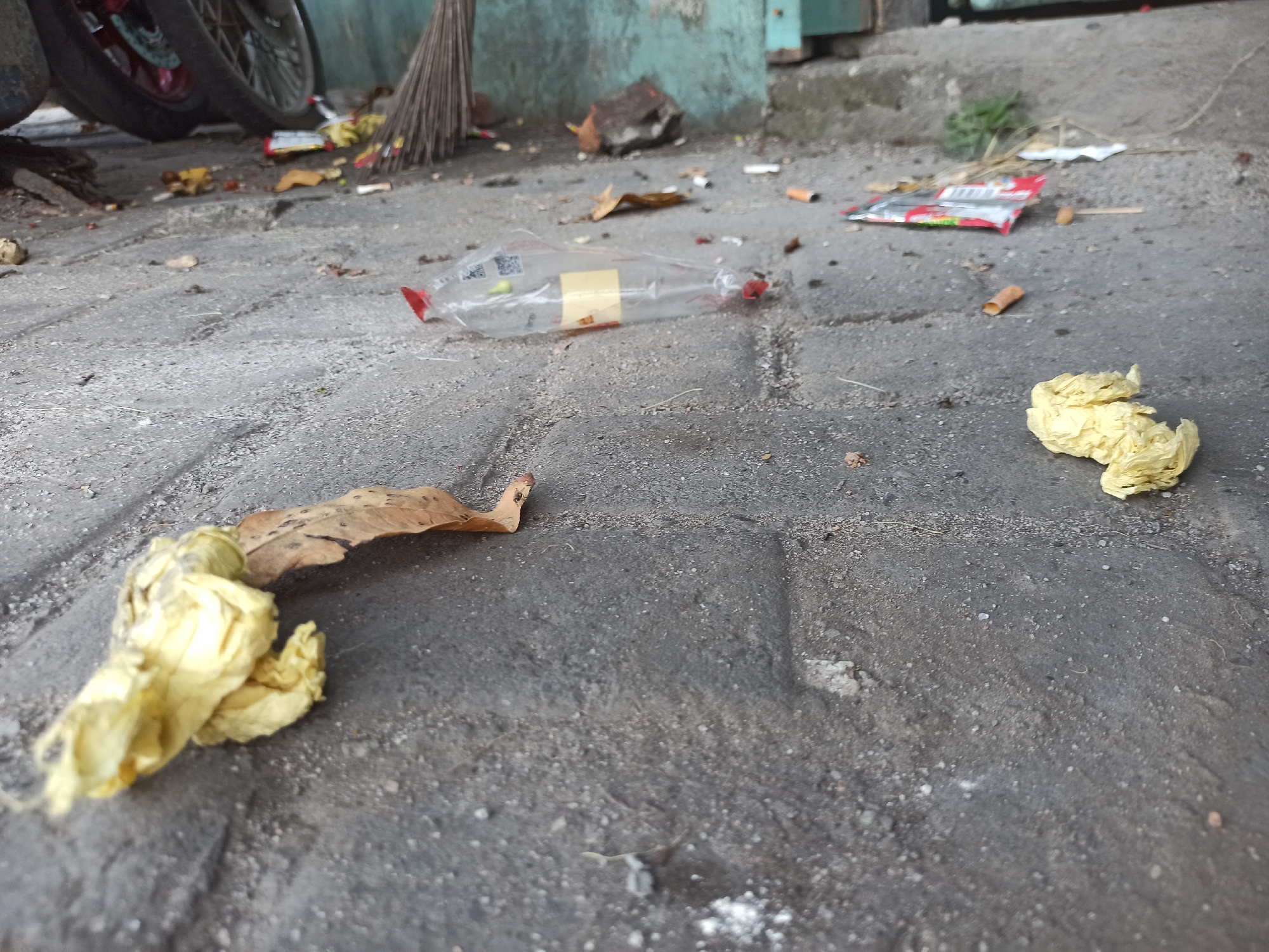 The image depicts a low-angle view of a dirty, paved city street in Medan, Indonesia. The ground is littered with debris, including cigarette butts, a plastic bottle, a crumpled yellow plastic wrapper, and a dry leaf. In the background, a broom leans against a weathered blue and green wall, and a portion of a motorcycle with red rims is visible on the left. The overall scene suggests a neglected urban environment.