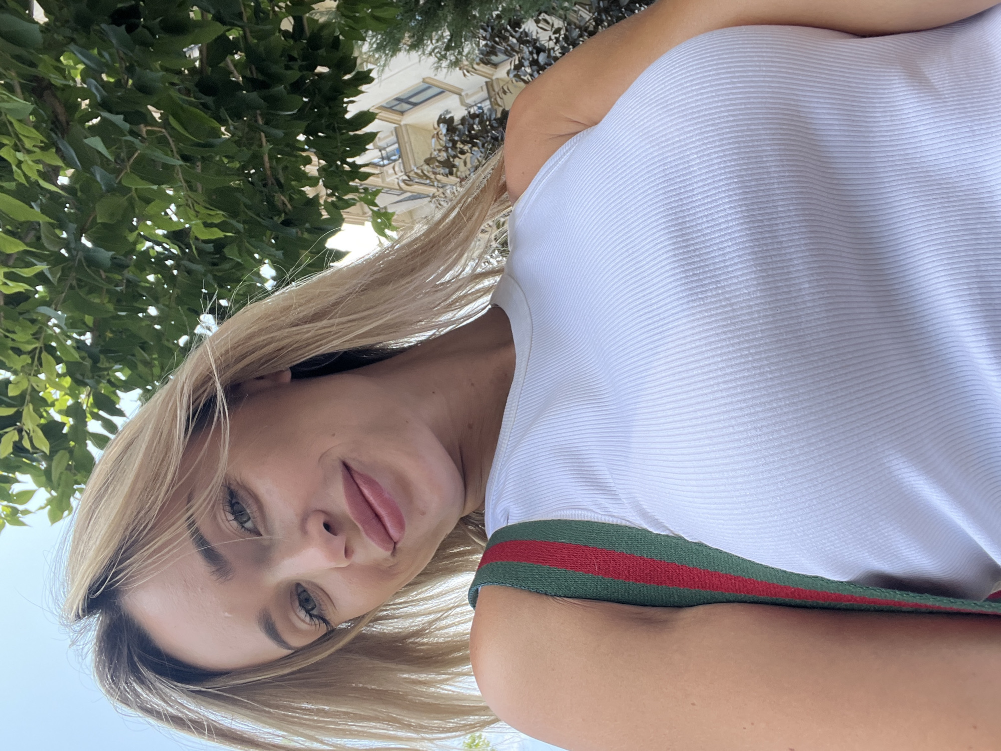 In Fontanka, Ukraine, a young woman with blonde hair and green eyes smiles at the camera while taking a selfie outdoors. She is wearing a white ribbed tank top and a bag with green and red stripes. Behind her, green foliage and the facade of a building are visible. The image is shot from a low angle, looking up towards the subject.