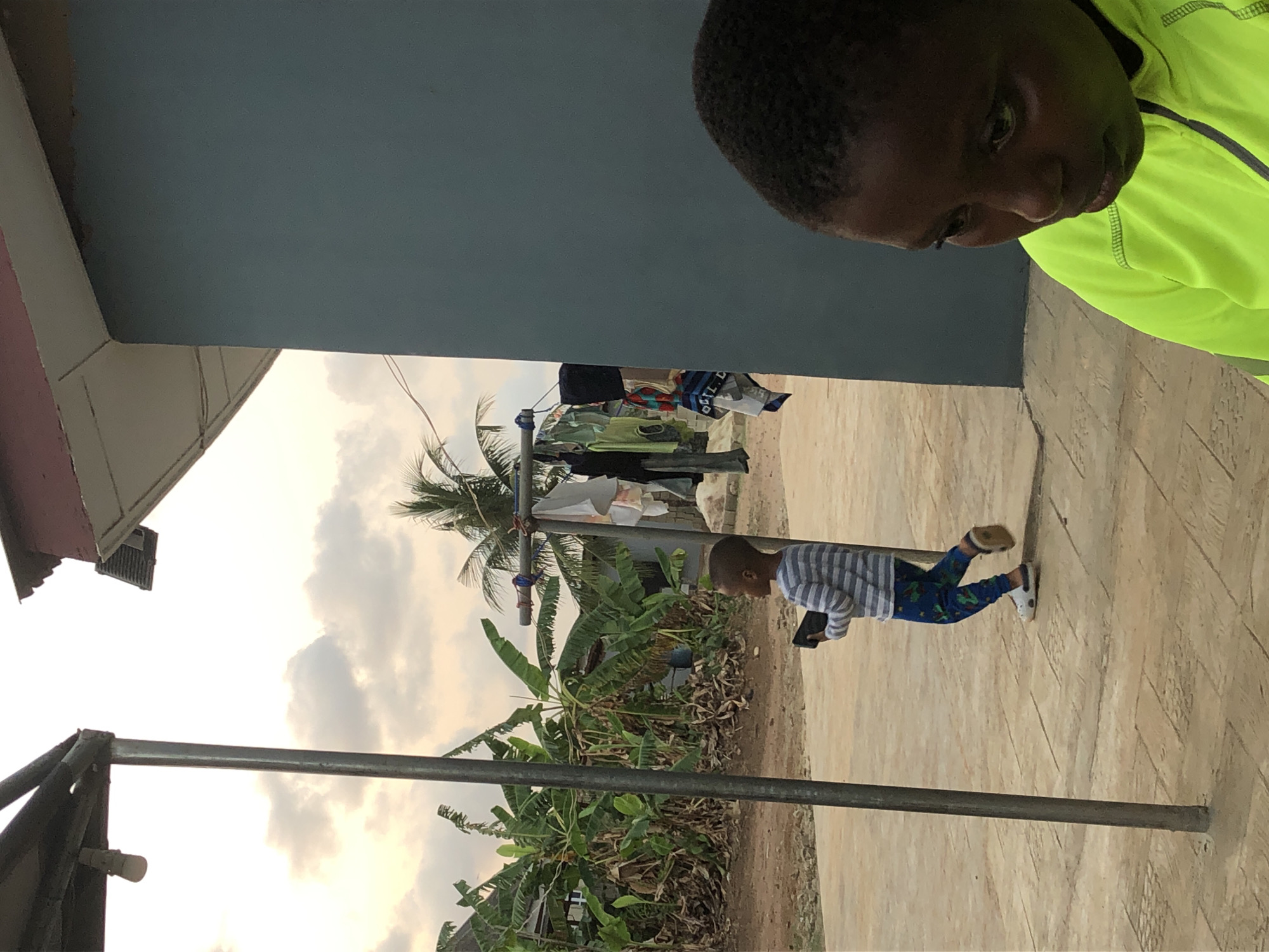 The image shows a courtyard in an outdoor setting, possibly in the Unknown, Unknown city. An adult, with dark hair and wearing a neon green and grey jacket, is partially visible in the upper right corner. A child in a horizontally striped top and printed pants is walking towards the right side of the frame, holding a dark object, possibly a phone or a small electronic device. In the background, a clothesline with laundry hanging on it is visible. Below the clothesline, there are banana trees and other plants. The courtyard has a brick-like paved surface. In the lower left corner, part of a structure with a metallic pole is seen. The sky is overcast with clouds.