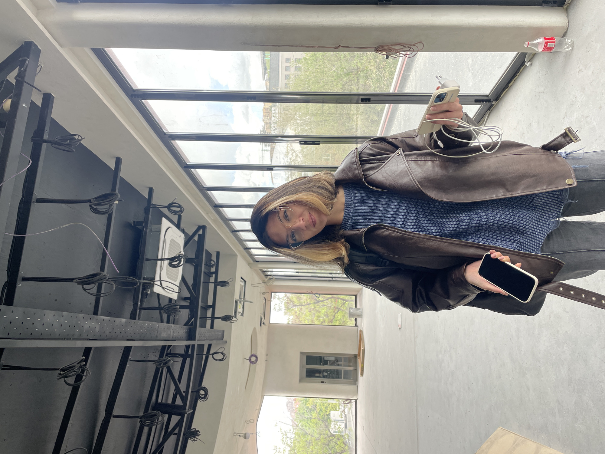 The photograph depicts a woman standing in what appears to be an interior space with a large skylight revealing an urban landscape. The woman, wearing a brown leather jacket and blue sweater, holds a smartphone in one hand and a charger in the other. The location is possibly in Yerevan, Armenia, suggested by the architectural style and urban layout visible through the skylight. The interior features exposed metal structures and wiring, possibly an industrial or studio setting. A Coca-Cola bottle sits near the skylight.
