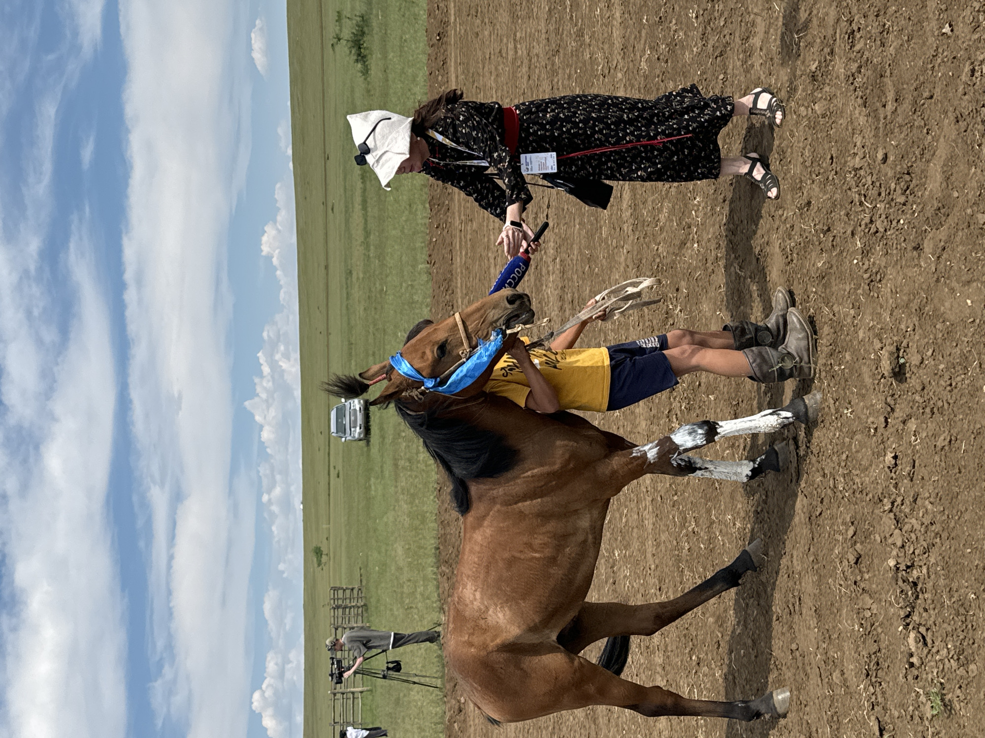 In Orongoi, Russia, a bay horse is being presented to a woman. The horse is decorated with a blue sash around its head and has white markings on its legs. The woman is wearing a black floral dress, a red sash, and a white hat with sunglasses. She is holding a microphone with "РОССИЯ" written on it, suggesting she is a reporter. A young person in a yellow shirt and blue shorts is standing next to the horse, holding its reins. In the background, a cameraman is filming, a fence, a car, and a grassy field with rolling hills can be seen under a partly cloudy sky. The ground in the foreground is dry and dusty.