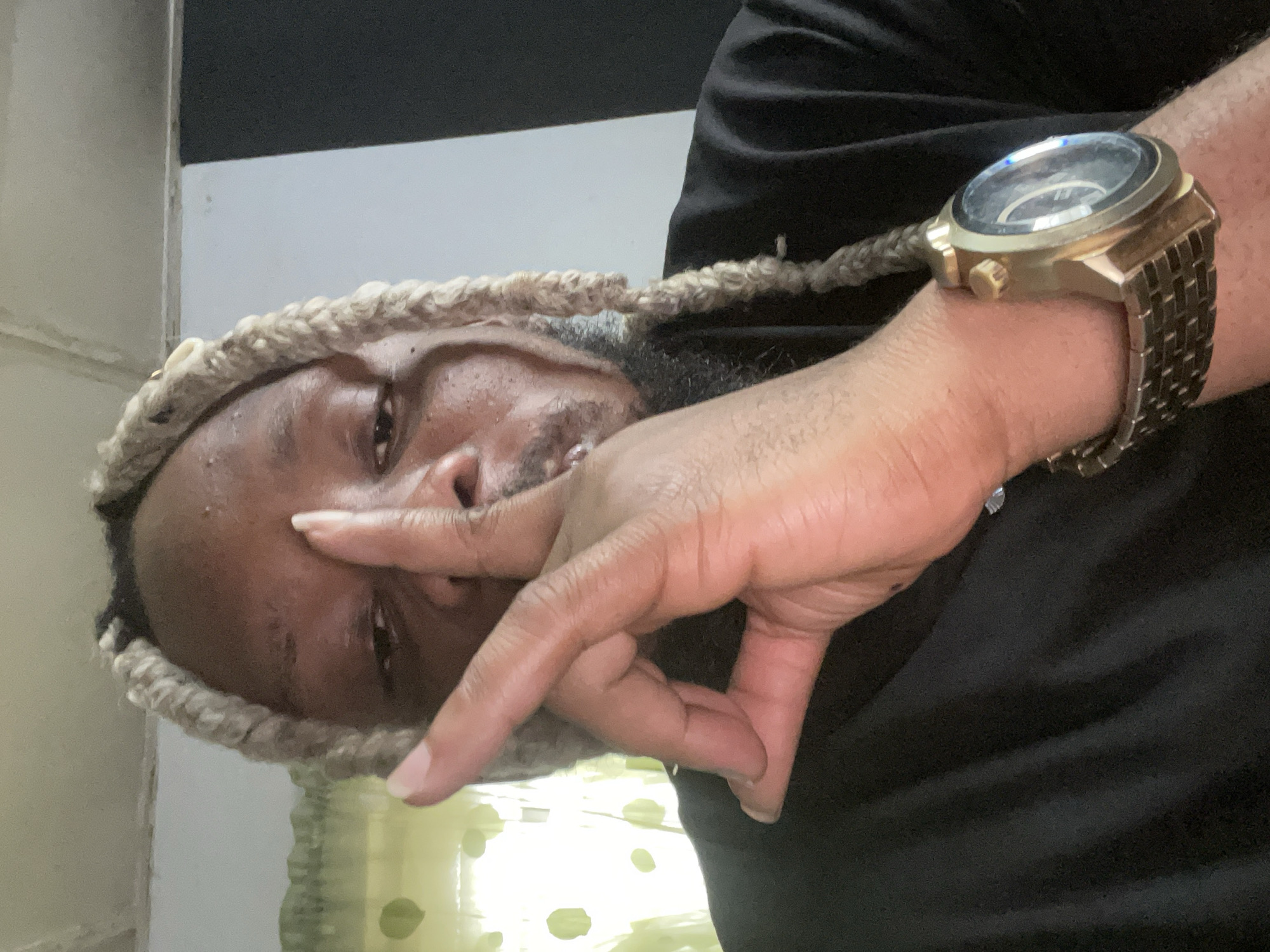 A close-up selfie features a man with blonde braided hair and a beard, wearing a black t-shirt. He is holding his right hand up to his forehead, with his index finger extended and touching his brow. His thumb and middle finger are bent, forming a shape that resembles the letter "L". A gold watch with a round face is visible on his left wrist. The background shows a dimly lit room with a window covered by a light green curtain with a dotted pattern. A black wall is visible to the right. The image does not provide any information to determine its location in the city of Jos, Nigeria.