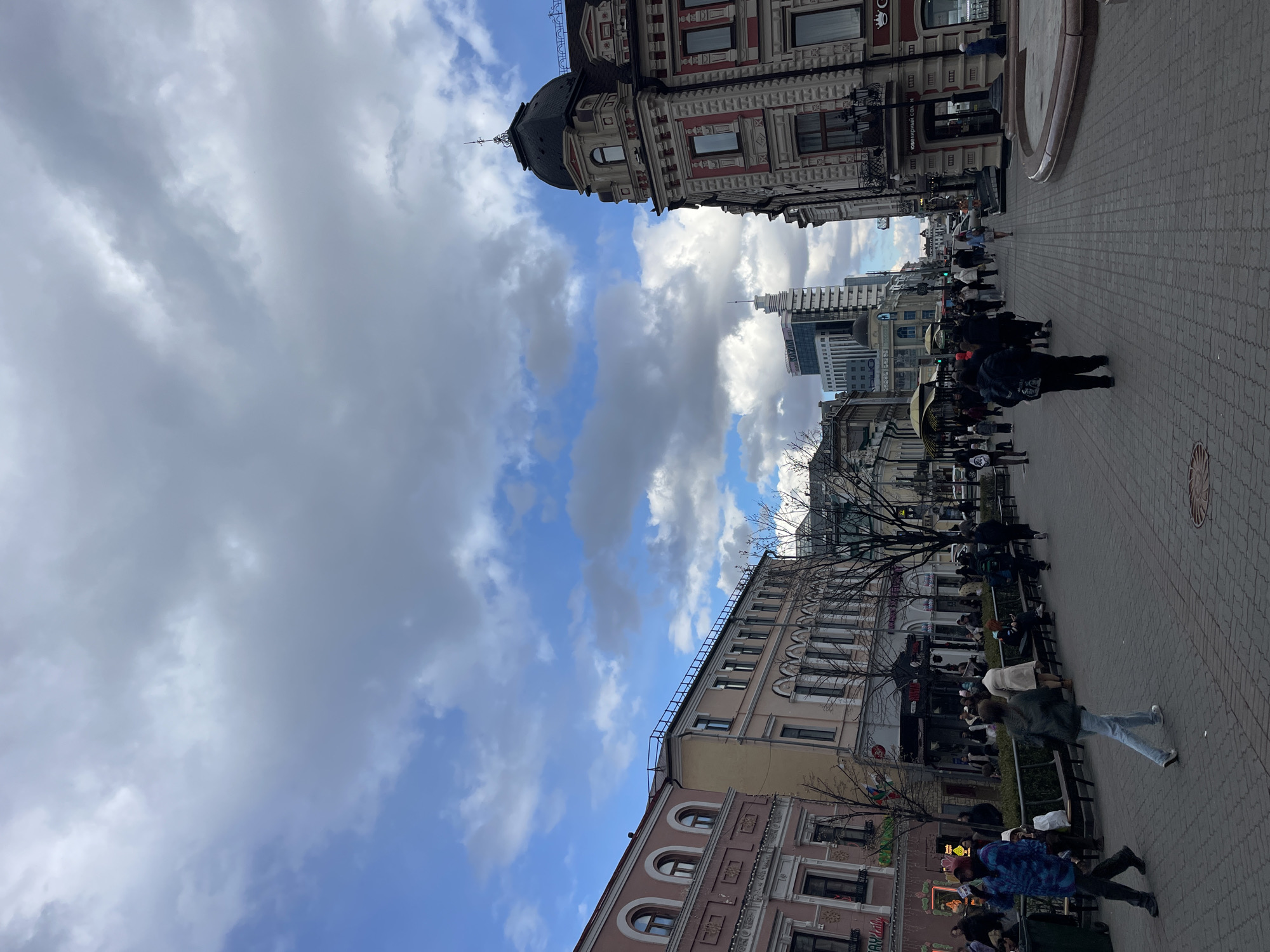 This wide-angle photograph captures a bustling city street in Kazan, Russia, under a partly cloudy sky. The image is presented rotated 90 degrees counter-clockwise, making the street appear to run vertically.
The scene is dominated by numerous pedestrians, both individuals and groups, strolling along the wide, brick-paved sidewalks. Some people are seated on benches or at outdoor establishments with umbrellas, suggesting a relaxed urban environment. Their attire is casual, fitting for a daytime city outing.
On the left side of the street (when viewed correctly), an ornate, multi-story building with a red and white facade and a prominent black-domed turret stands out, showcasing historic architecture. Other older buildings with detailed windows and decorative elements line this side of the street. In contrast, taller, more modern buildings with glass facades are visible further down on the right, indicating a blend of old and new urban development.
The sky features patches of bright blue interspersed with large, fluffy white and grey clouds. The presence of bare tree branches suggests the season is likely late autumn, winter, or early spring. Street furniture such as lamps, benches, and planter boxes are visible along the pedestrian paths. A clear sign on one of the buildings displays the Russian text "ЮВЕЛИРНЫЙ СОН," which translates to "JEWELRY DREAM" or "JEWELRY SLEEP," serving as a key location cue. The consistent daylight indicates the time of day is likely mid-morning to early afternoon. The overall atmosphere is one of a lively and active city center.