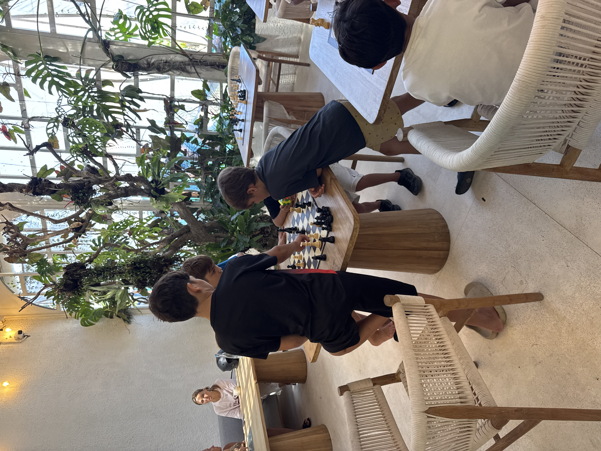 The image shows a brightly lit indoor area, likely a café or restaurant, possibly in North Kuta, Indonesia, given the tropical plants visible. Three boys are engaged in playing chess at a wooden table. One boy is wearing a black t-shirt and shorts, another is wearing a grey t-shirt and shorts, and a third boy is wearing a white t-shirt and shorts. The chess set is placed on the table between them. The furniture appears to be wooden with woven white rope chairs. In the background, a woman sits at a table. Abundant tropical foliage is visible, growing near large windows or an open-air structure, suggesting a tropical climate.