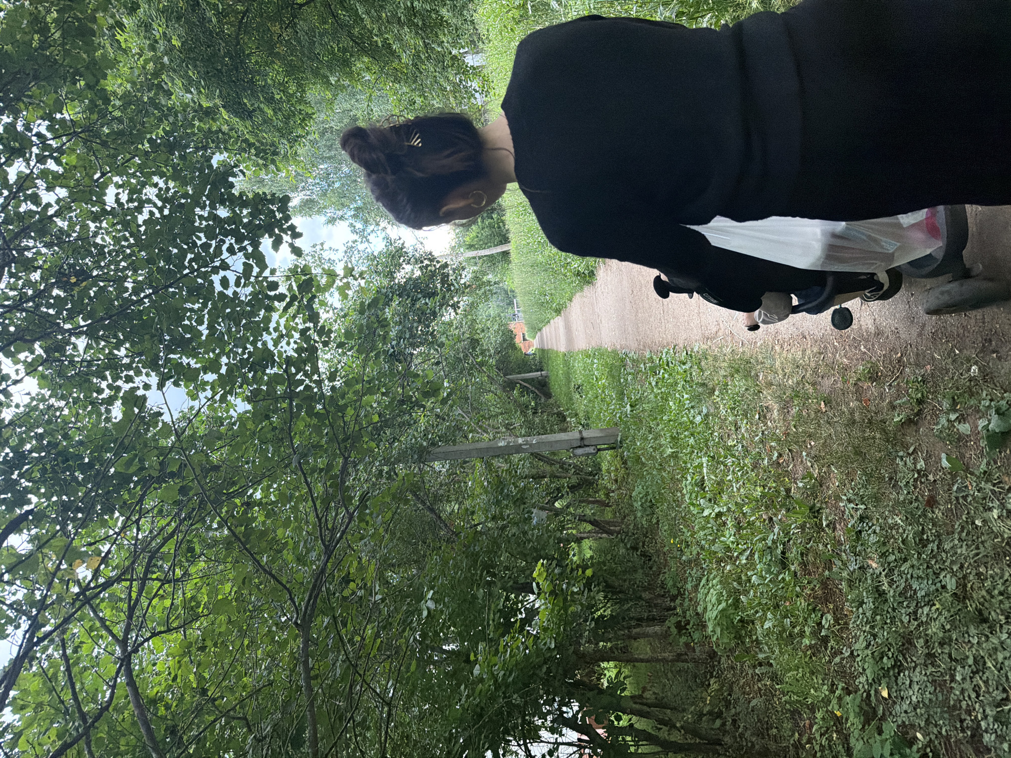 A person wearing a black outfit is seen from behind, walking down a path lined with green foliage. They have their hair pulled up in a bun and are wearing gold hoop earrings. The path appears to be made of dirt or gravel and is surrounded by trees and tall grass, suggesting a natural or park-like setting. The person is pushing a stroller that has a white bag in it. The scene is outdoors and appears to be during the daytime, with natural light filtering through the leaves. The overall mood is one of peacefulness and possibly a leisurely stroll through nature. There are no visible text elements in the image.