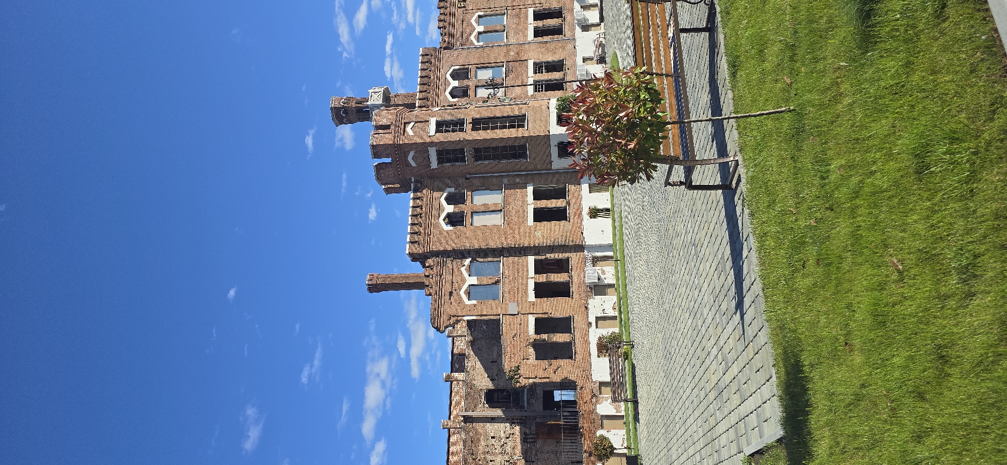The image shows a tall, brick building in a state of disrepair, possibly an old factory or industrial building. The building has multiple stories and many empty window openings. The architecture includes decorative elements such as corbels and crenellations. The building is partially covered in vegetation and appears to be historical.
To the right of the building is a paved walkway made of interlocking pavers. Adjacent to the walkway is a strip of green lawn. A small tree and a bench are located near the walkway. The sky above is blue with some scattered clouds.
The location is Unknown, Unknown.