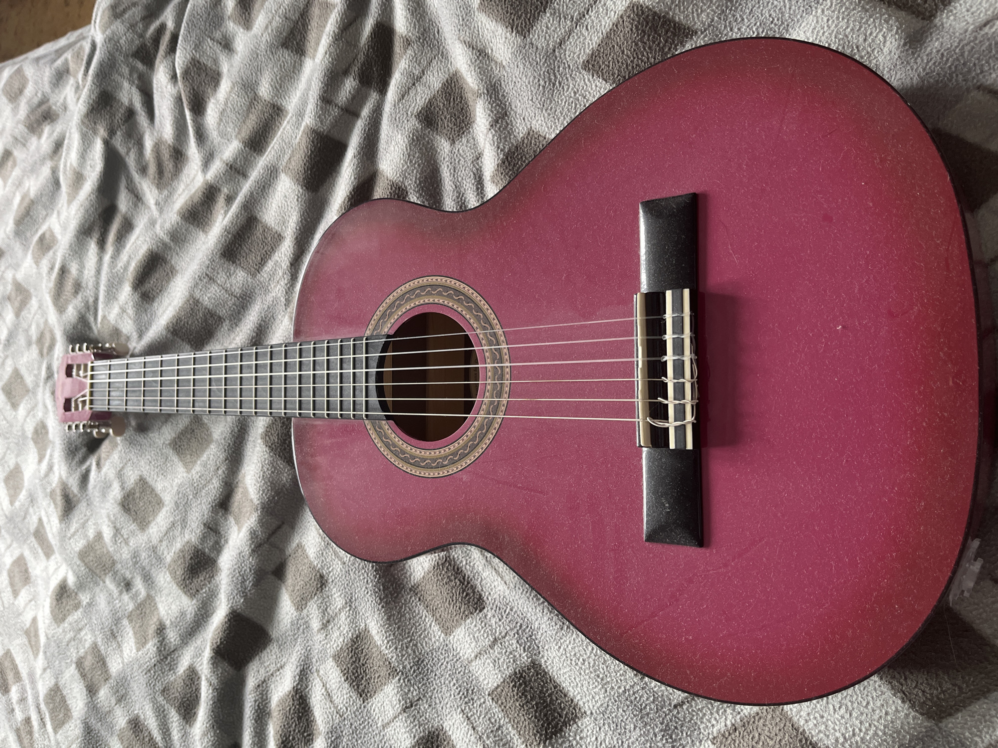 An acoustic guitar with a reddish-pink body lies on a grey and brown checkered blanket. The guitar is positioned vertically, with the headstock at the top of the image. The fretboard and strings are clearly visible, as is the soundhole with its decorative rosette. The body of the guitar has a matte finish and appears to be dusty. The blanket has a soft, textured surface. The lighting suggests an indoor setting. The image was taken in Moscow, Russia.