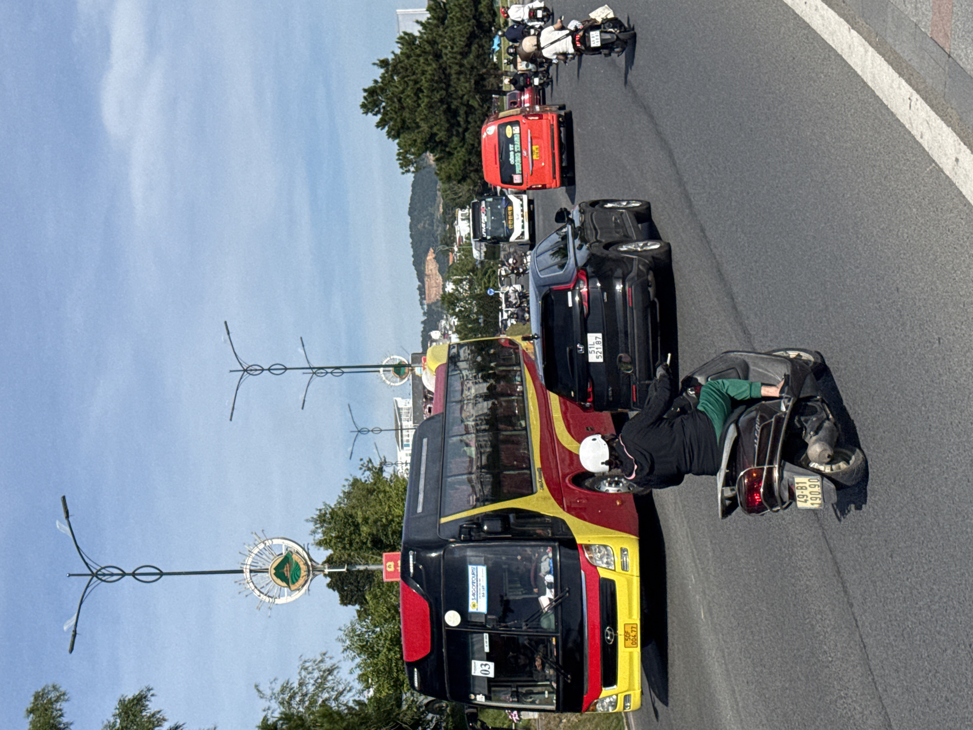 The image depicts a street scene in Da Lat, Vietnam, on a sunny day with a clear blue sky and scattered clouds. The foreground features a dark-colored scooter with a license plate reading "49-B1 190.90" and a rider wearing a dark jacket and green pants with a white helmet. To the left and slightly behind the scooter is a large, yellow and red bus with the number "03" on its front and a "SAIGONTOURS" logo. The bus is traveling on a multi-lane asphalt road. Several other vehicles, including cars and motorcycles, are visible further down the road. In the background, trees and buildings line the street, with a hilly landscape in the distance. Two ornate lampposts with decorative circular emblems are prominent on either side of the road. One emblem on the left features a green landscape with a mountain and a sunburst. The overall impression is of a busy urban environment in Da Lat.