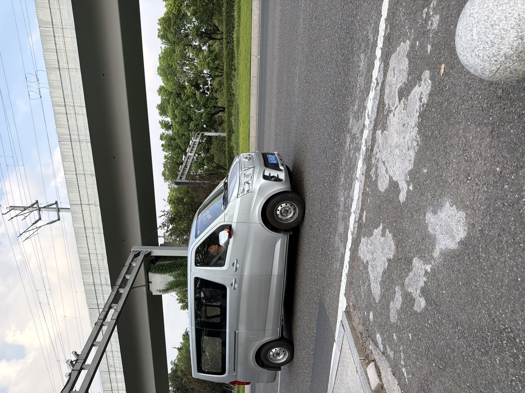 A silver minivan is driving on a paved road in Jiangjin, China, under an elevated structure with train tracks above. The minivan's license plate is partially visible and reads "JIA B 355P". A person is driving the minivan, and their face is visible through the driver's side window. The road has a white line marking the lane. In the background, there are trees and green grass. A concrete barrier is visible along the side of the road, and a granite sphere is on the right side of the image. The sky is cloudy.