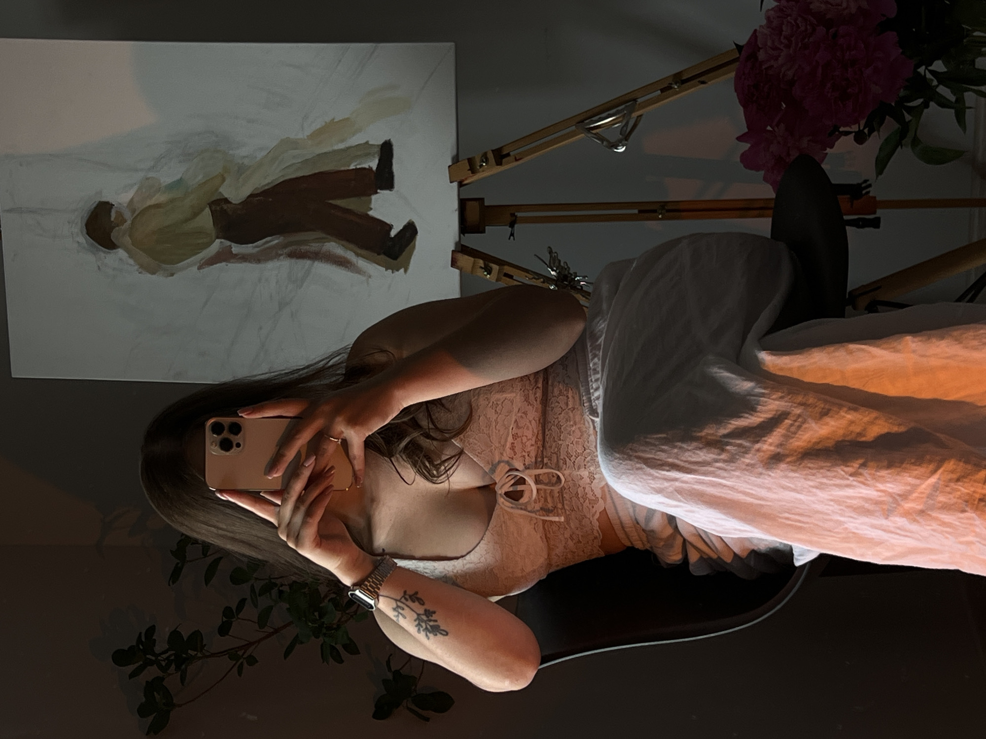The image shows a young woman sitting on a chair in what appears to be an artist's studio or a room with art supplies. She is taking a selfie with her phone, which is held up to her face, obscuring her features. The woman is wearing a light-colored, lacy top and a long, white skirt. A silver wristwatch is visible on her left wrist, and she has a tattoo on her left arm.
Behind her, an easel stands with an unfinished painting of two figures. The painting is in a loose, abstract style, with muted colors. To the left of the woman, a plant with dark green leaves is visible, and to the right, a vase of vibrant pink flowers adds a pop of color to the scene. The room is dimly lit, with warm light casting shadows and highlighting the textures of the fabric and the painting. The overall mood is intimate and creative.
The provided information states the location is Ternopil, Ukraine. However, there are no specific visual cues in the image that would confirm this location. The scene is generic enough to be in many urban or suburban environments.