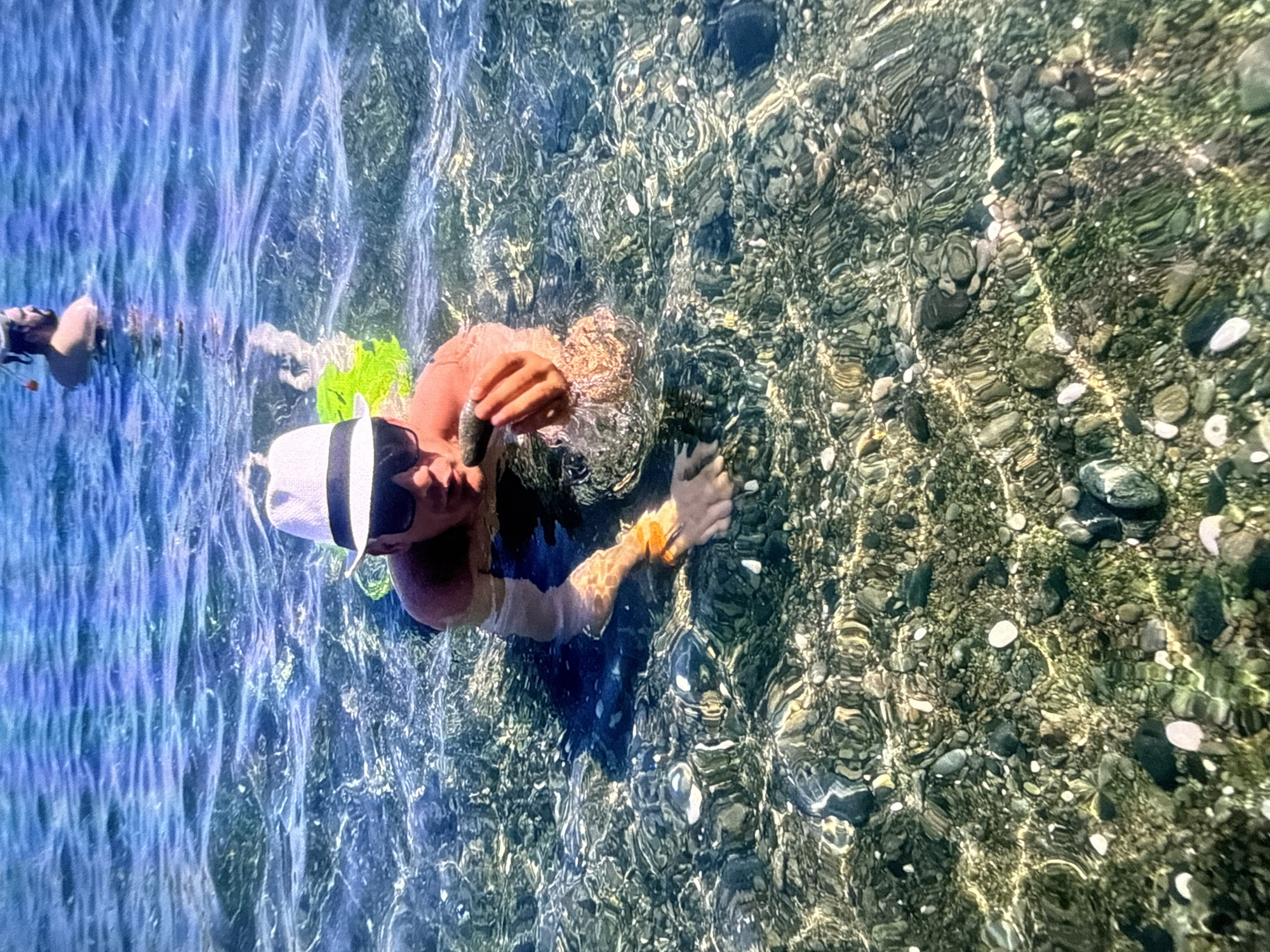 In a clear, shallow body of water with a pebbled seabed, a man wearing a white fedora and sunglasses is partially submerged. He is holding a dark, oblong object in his right hand. His left arm is extended, with an orange bracelet on his wrist. Another person, wearing a snorkel mask, is visible further in the background. The water is illuminated by sunlight, creating dappled patterns on the seabed. The scene appears to be a recreational setting, possibly a beach or a cove. The location is not Szczecin, Poland, as there are no elements in the image to suggest this specific city.