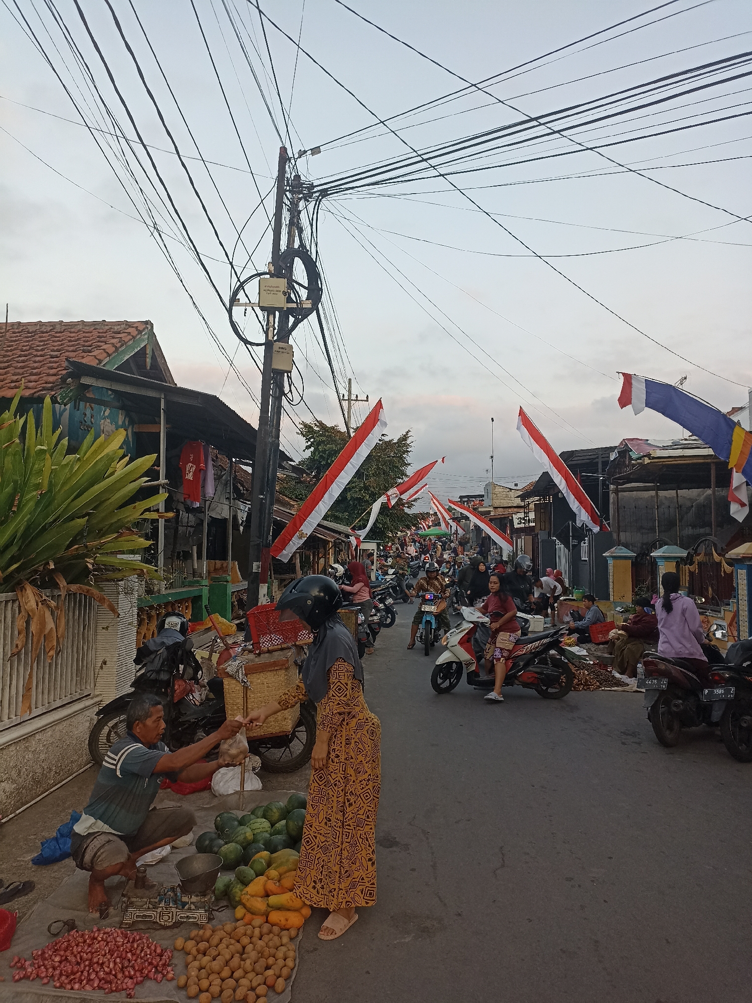 A busy street scene in Mojopanggung, Indonesia, is captured at dusk. The road is filled with motorcycles, some ridden by people, others parked. Red and white flags are strung across the street, a common sight during Indonesian Independence Day celebrations. On the left side of the road, a vendor is selling produce from his stall. He is squatting behind a scale and has piles of red onions, potatoes, watermelons, and papayas laid out on a tarp. A woman in a patterned dress is interacting with the vendor, possibly making a purchase. Shops and houses line the street, their facades weathered. Overhead, a dense network of electrical wires crisscrosses the sky, illuminated by the fading light. The atmosphere appears lively, with a mix of commerce and community gathering.