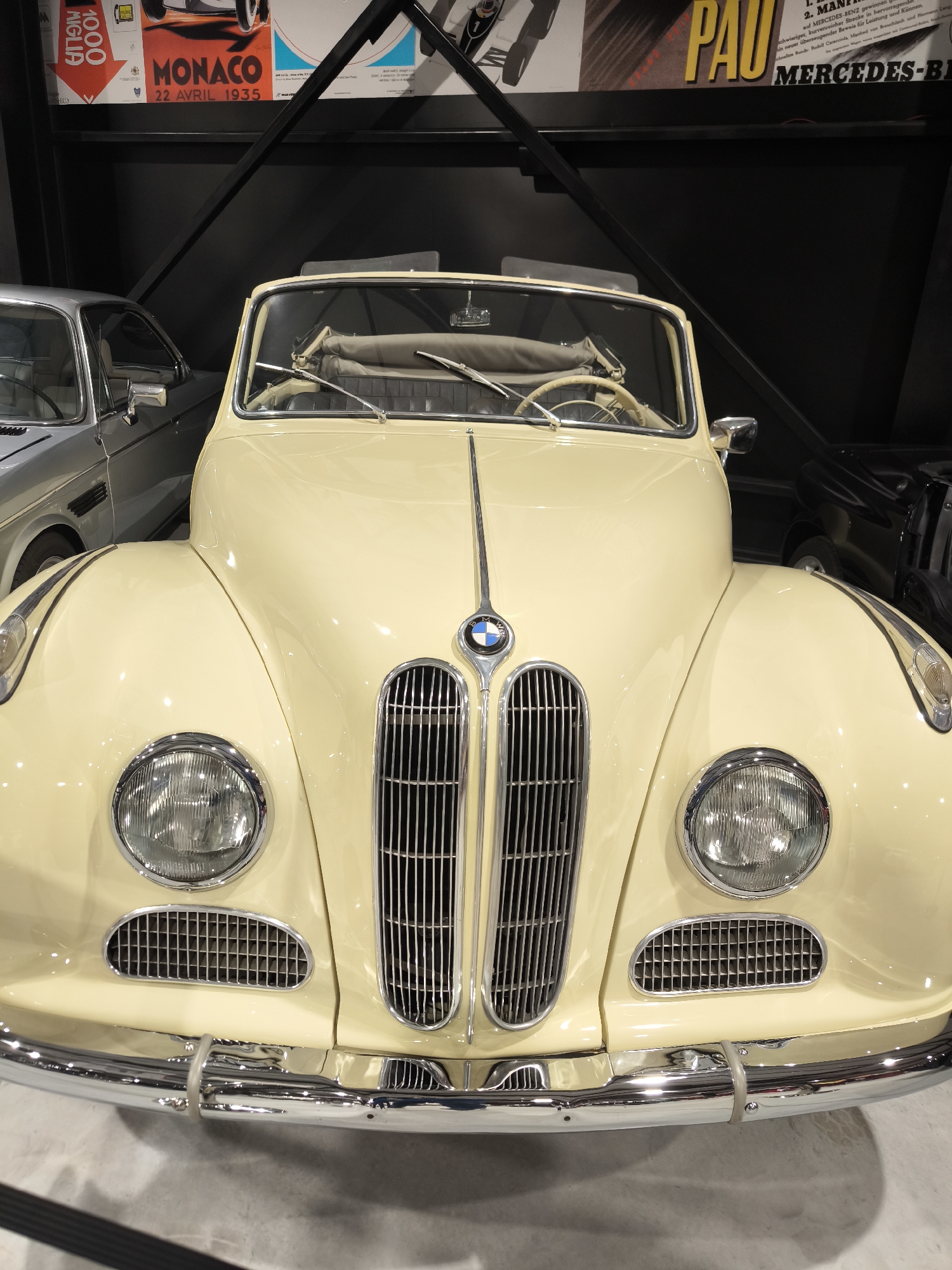 The image is a close-up of the front of a classic, cream-colored BMW convertible. The car has prominent headlights, a tall chrome grill with the BMW logo, and a shiny chrome bumper. The car's interior features a light-colored steering wheel and what appears to be a stowed convertible top. Behind the featured car, partially visible, are two other cars – one silver and one black.
The background suggests an indoor exhibition or museum setting. There are black metal support beams visible, along with posters on the wall featuring vintage racing advertisements. One poster mentions "Monaco, 22 Avril 1935" and another prominently features the word "PAU" above the name "MERCEDES-BENZ."
The information about Kapatovo, Bulgaria, cannot be verified in this image.