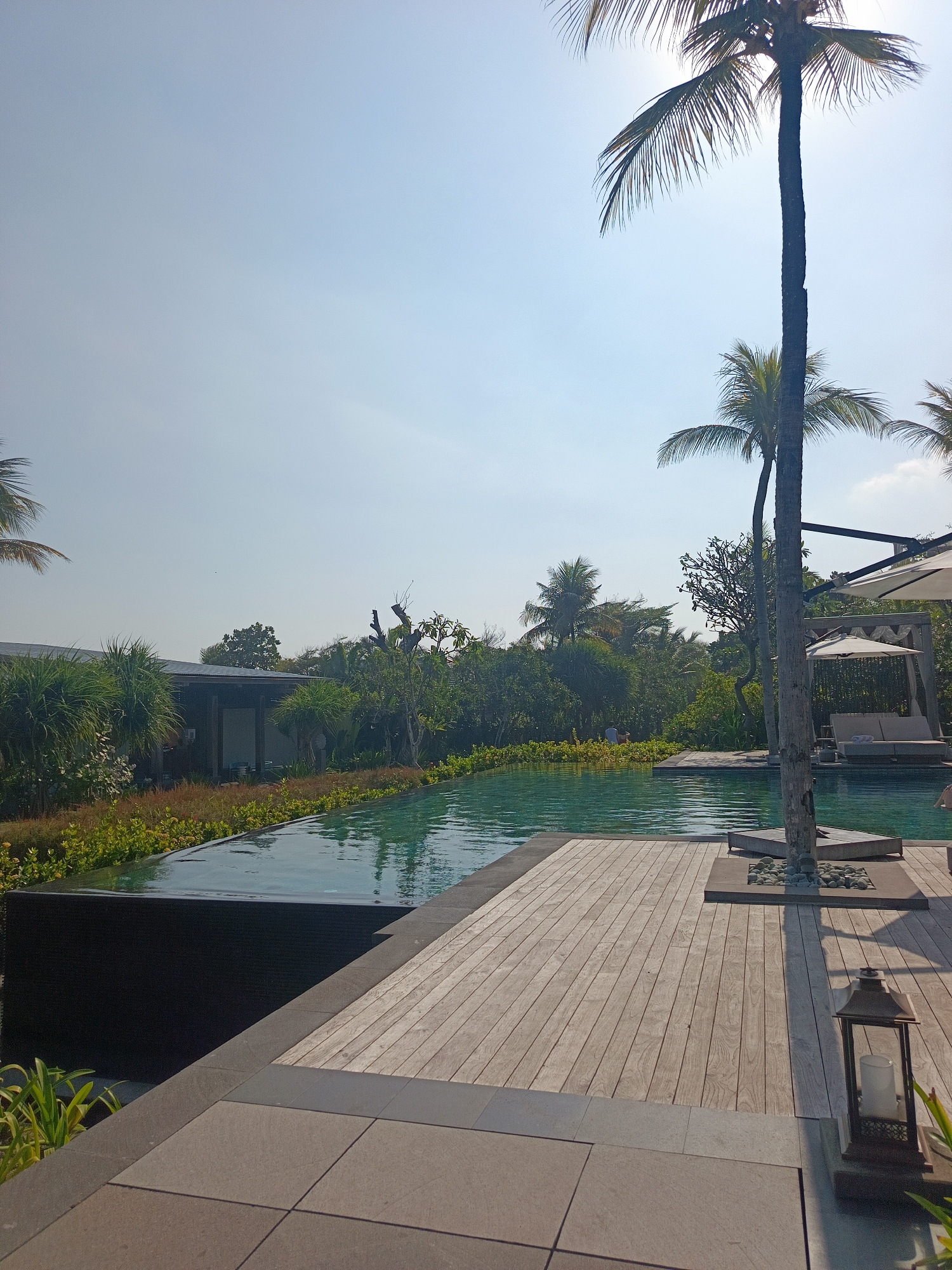 A bright, sunny day in North Kuta, Indonesia, as evidenced by the clear blue sky. The scene depicts an infinity pool with calm, turquoise water. The poolside is furnished with a wooden deck and a shaded cabana with lounge chairs. Lush greenery, including palm trees and various tropical plants, surrounds the pool area, suggesting a resort or private villa setting. A decorative lantern stands on the wooden deck, and some scattered pebbles are visible around a tree trunk. The architecture visible in the background appears modern with a dark-tiled roof and open-air structures. There are a few people visible in the distance, near the pool and the cabana, engaged in leisure activities.