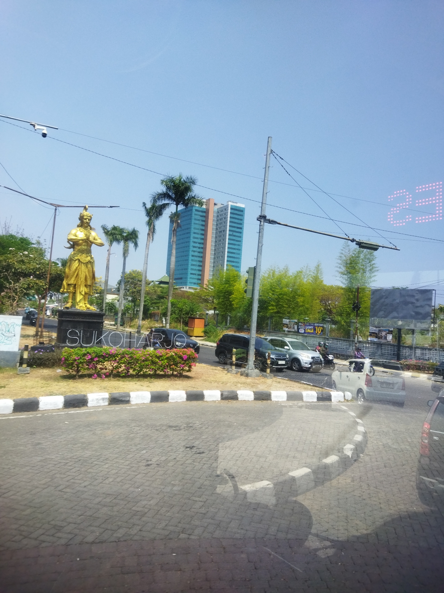 The image captures a street scene in Grogol, Indonesia, under a clear blue sky. In the foreground, a paved road curves to the right, bordered by a curb with alternating black and white segments. To the left of the road is a grassy area with a golden statue of a person standing on a pedestal. The pedestal is inscribed with the word "SUKOHARJO." A line of flowering bushes with pink blooms runs in front of the pedestal. In the background, several tall palm trees and other greenery are visible. Behind the trees, two modern buildings, one tall and blue with many windows, and another shorter and orange, stand out against the sky. A street light and traffic signals are also present, along with several cars and a motorcycle on the road. A digital display showing "23" in pink lights is visible in the upper right corner of the image. The overall scene depicts a city intersection with public art, signage, and vehicular traffic.