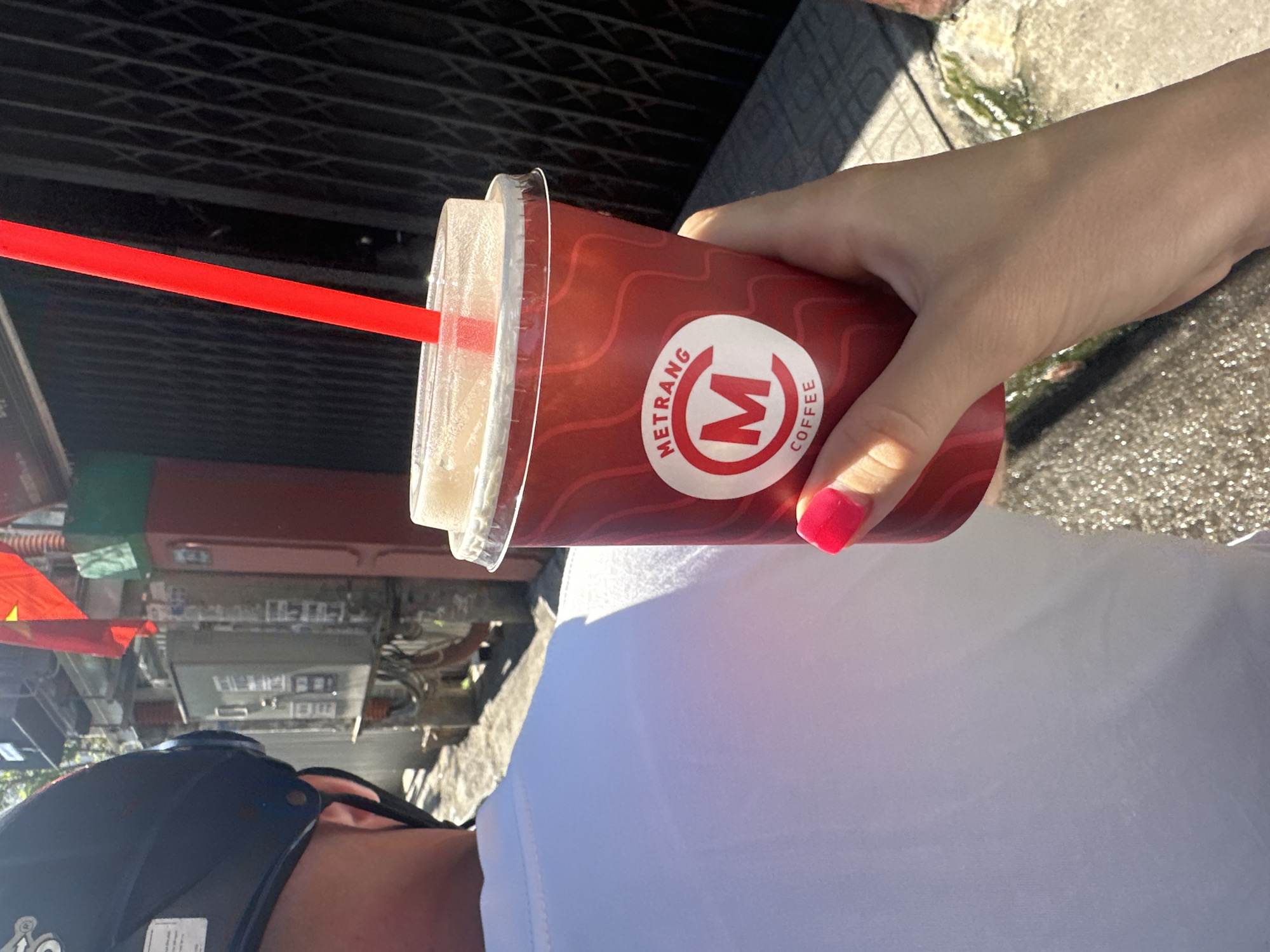 A person wearing a black helmet and a white shirt holds a "METRANG COFFEE" beverage in their right hand, with a red straw protruding from the lid. The fingernail of the index finger has red polish. The location is identified as Bac Nha Trang Ward, Vietnam, based on signage and flags visible in the background, and the general urban environment. Electrical boxes and other infrastructure are visible, suggesting a street scene. The person is likely taking a selfie while outdoors.