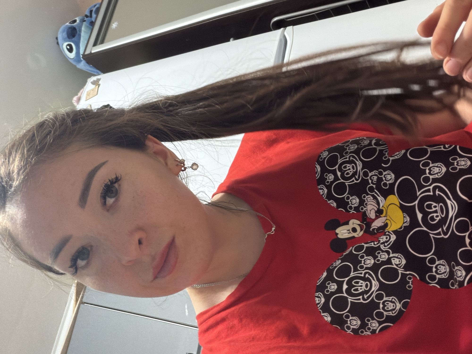 This is a vertically oriented, eye-level, close-up selfie of a young woman with long, dark brown hair. She is wearing a red t-shirt with a black and white Mickey Mouse pattern. Her hair is pulled back and extends upwards, with her left hand holding a section of it. She has light skin with freckles, prominent eyelashes, and is wearing light makeup. A thin silver necklace is visible around her neck. In the upper left corner of the image, a blue stuffed toy with large eyes can be seen. The background appears to be indoors, with a white surface and what looks like a cabinet or shelf behind her. The lighting suggests it is daytime. The context provided indicates the location is Tujmazy, Russia.
