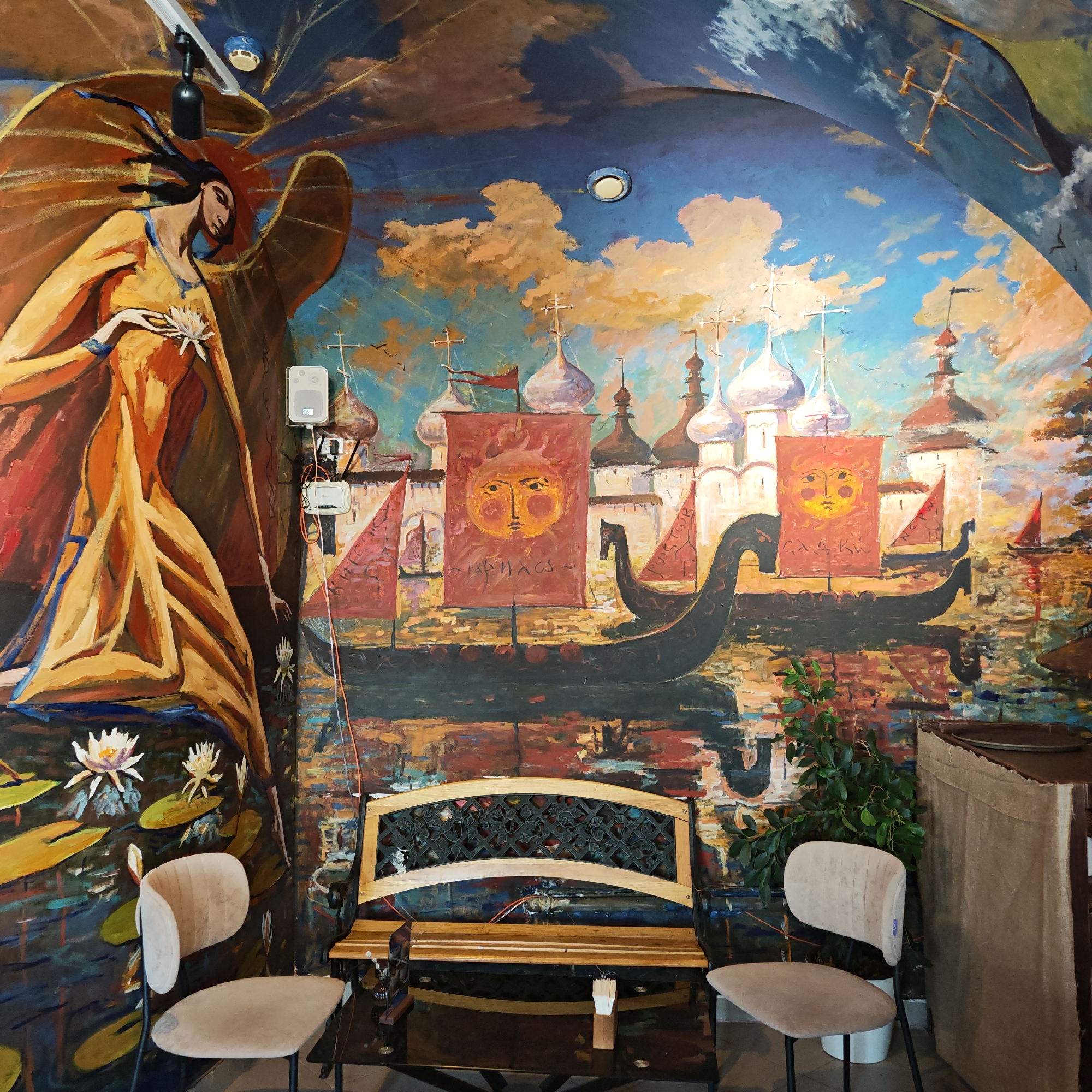 The image captures an indoor space in an unknown city. The room's primary feature is a large, colorful mural covering the walls and ceiling, depicting a fantastical scene with elements of Russian folklore and art. A central female figure with wings and a flower stands on the left, near water lilies. The background displays a cityscape with churches, ships with sun-emblazoned flags, and a vibrant sky. Below the mural, there's seating consisting of two chairs and a bench. A dark glass table sits in front of the bench. A potted plant is placed near one of the chairs, and electrical boxes are visible near the mural on the left.