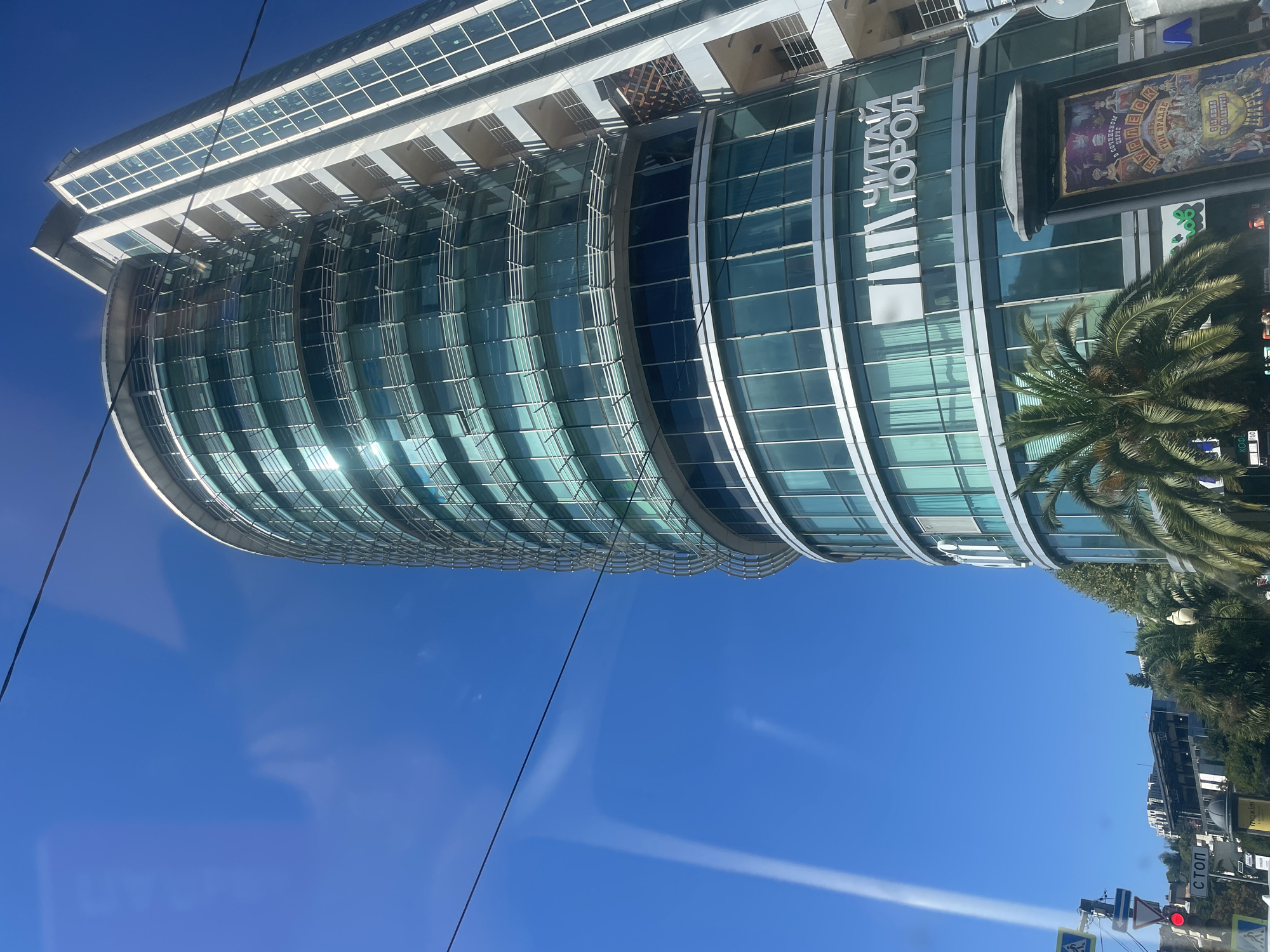 This is a daytime, eye-level, slightly angled, outdoor photograph of a modern, multi-story building with a curved glass facade and a rectangular section on the right, located in Sochi, Russia. The building has a prominent "ЧИТАЙ ГОРОД" (Read City) logo in white letters on its glass surface. In the foreground, a palm tree is visible, and to the left, traffic lights and pedestrian crossing signs are present. A large poster or advertisement with colorful imagery is also visible in the lower right portion of the frame, next to the palm tree. The sky is clear and blue.