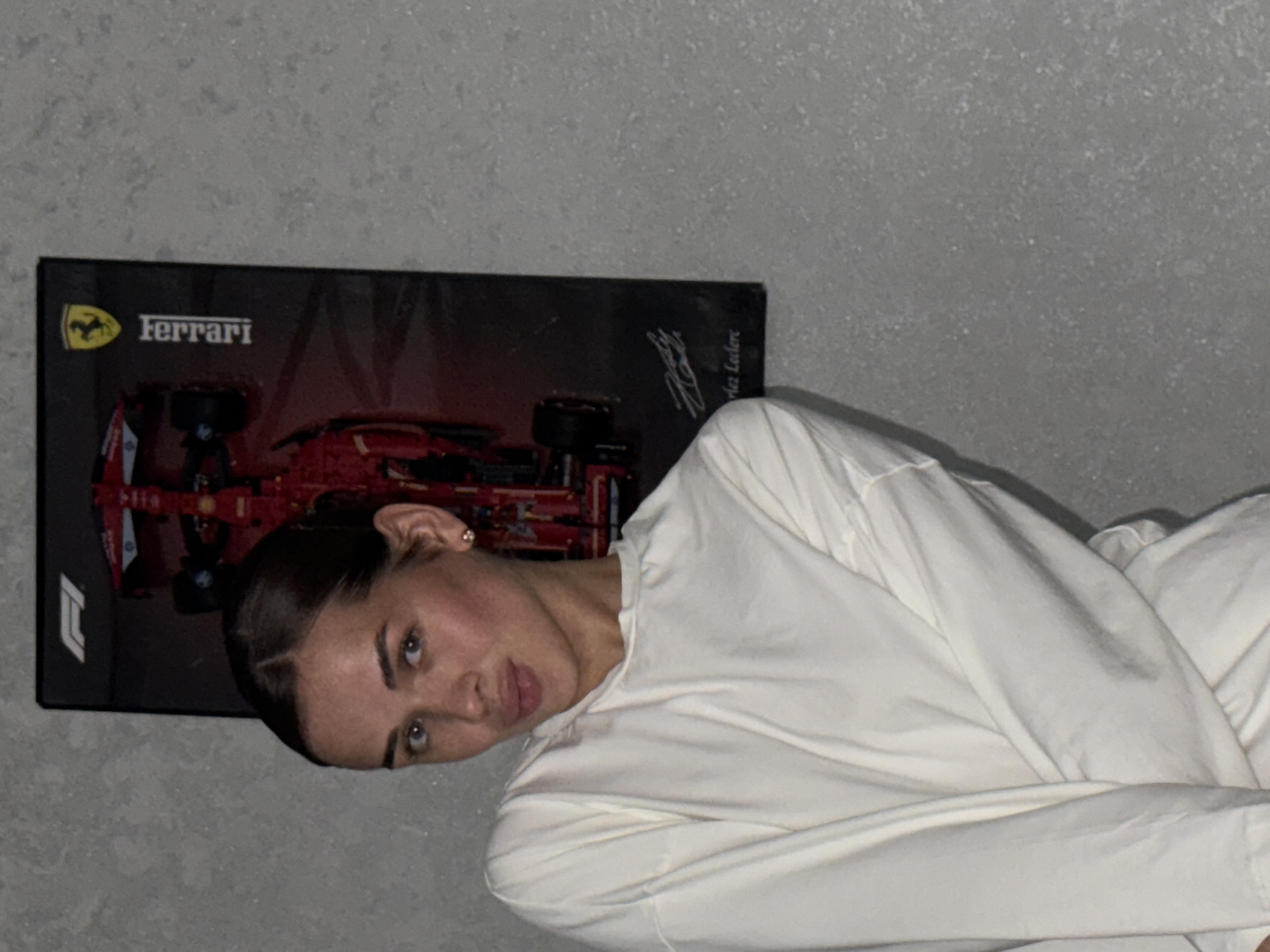 A woman lies in bed in a room in what appears to be Лобаново, Russia. She is wearing a white long-sleeved shirt. Behind her is a poster of a Ferrari race car, featuring the Ferrari logo and the signature of Charles Leclerc. The background wall is light gray.
