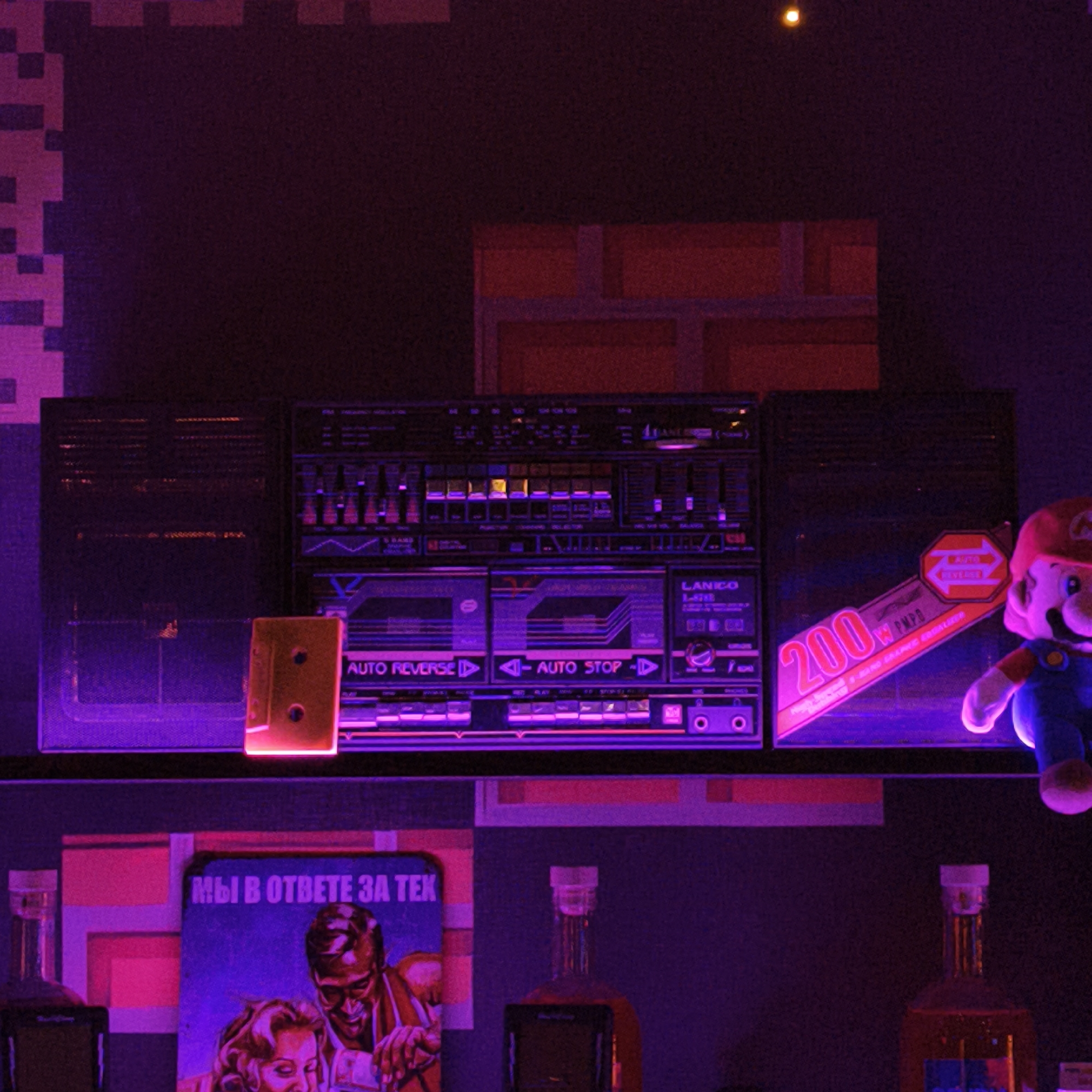 A dimly lit, retro-themed scene shows a shelf with various objects against a backdrop of pixelated patterns reminiscent of classic video games. Dominating the shelf is a large, vintage LANICO L-200 boombox with a cassette inserted, illuminated by neon-like purple and pink lights. A plush Mario figure stands to the right of the boombox. Below, a book titled "MЫ В ОТВЕТЕ ЗА ТЕК" with a graphic design featuring a man and a woman, is visible alongside several bottles, possibly alcoholic beverages. Based on the details and clues, the location is identified as Hrodna, Belarus, given the Cyrillic book title, which means "We are responsible for the text".