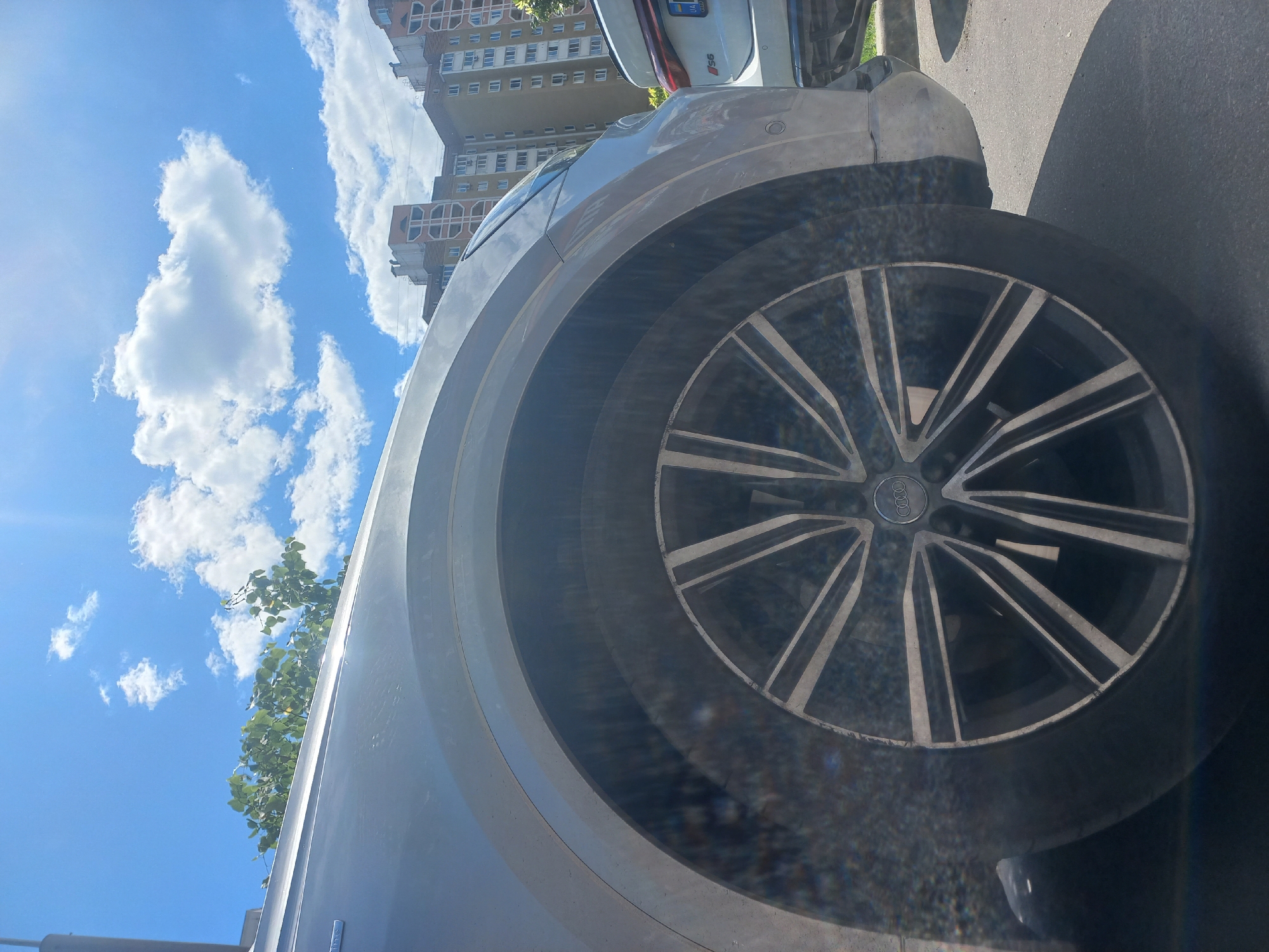 The image displays the front-left side of a light-colored Audi car, likely silver or light gray, parked outdoors under a bright blue sky. The car's prominent multi-spoke wheel is a focal point, featuring a two-tone design of silver and dark gray spokes, with the Audi rings logo distinctly visible on its center cap.
In the background, a tall, light-colored residential building with numerous windows and balconies stands against the sky. Partially visible behind the primary Audi is the rear of a white car, identified by an "RS6" badge and a license plate bearing the letters "UA", suggesting a Ukrainian setting. The sky is clear with scattered white, fluffy clouds. Green foliage from a tree is visible in the upper left section of the frame. The bright sunlight indicates it is daytime. The ground appears to be asphalt paving. No people or specific activities are depicted; the scene is static, focusing on the parked cars and their urban surroundings.