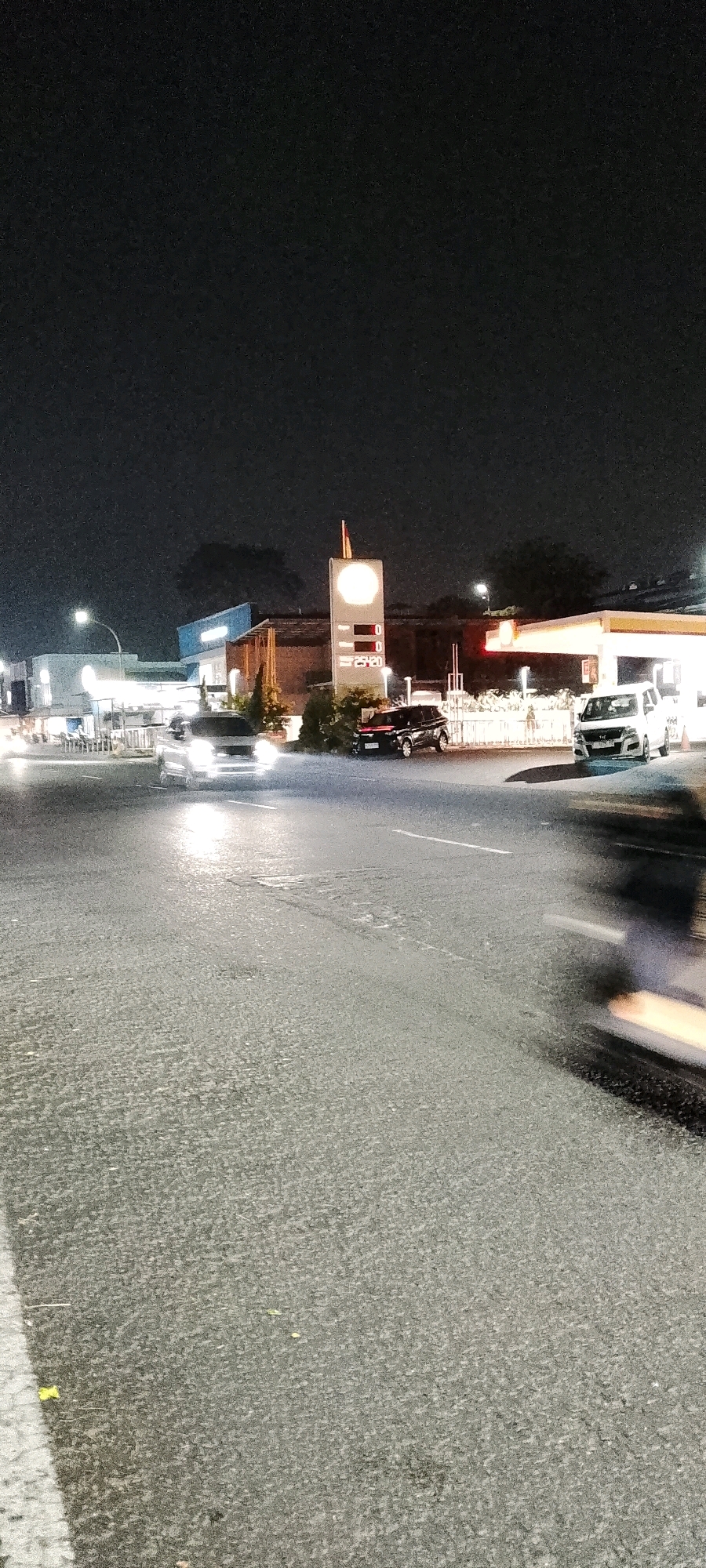 The image captures a night scene at a gas station in Cileungsi, Indonesia. The foreground shows a wet asphalt road with tire marks and a white line on the left side. In the midground, several cars are visible: a silver car with its headlights on is approaching from the left, a black SUV is parked at the gas station, and a white SUV is also at the gas station. A large gas station sign with a circular illuminated display and numerical prices is prominent, indicating it is operational. The background features the gas station canopy, buildings, and trees under a dark sky, suggesting an urban or suburban setting. The overall lighting suggests artificial illumination from the gas station and streetlights.
