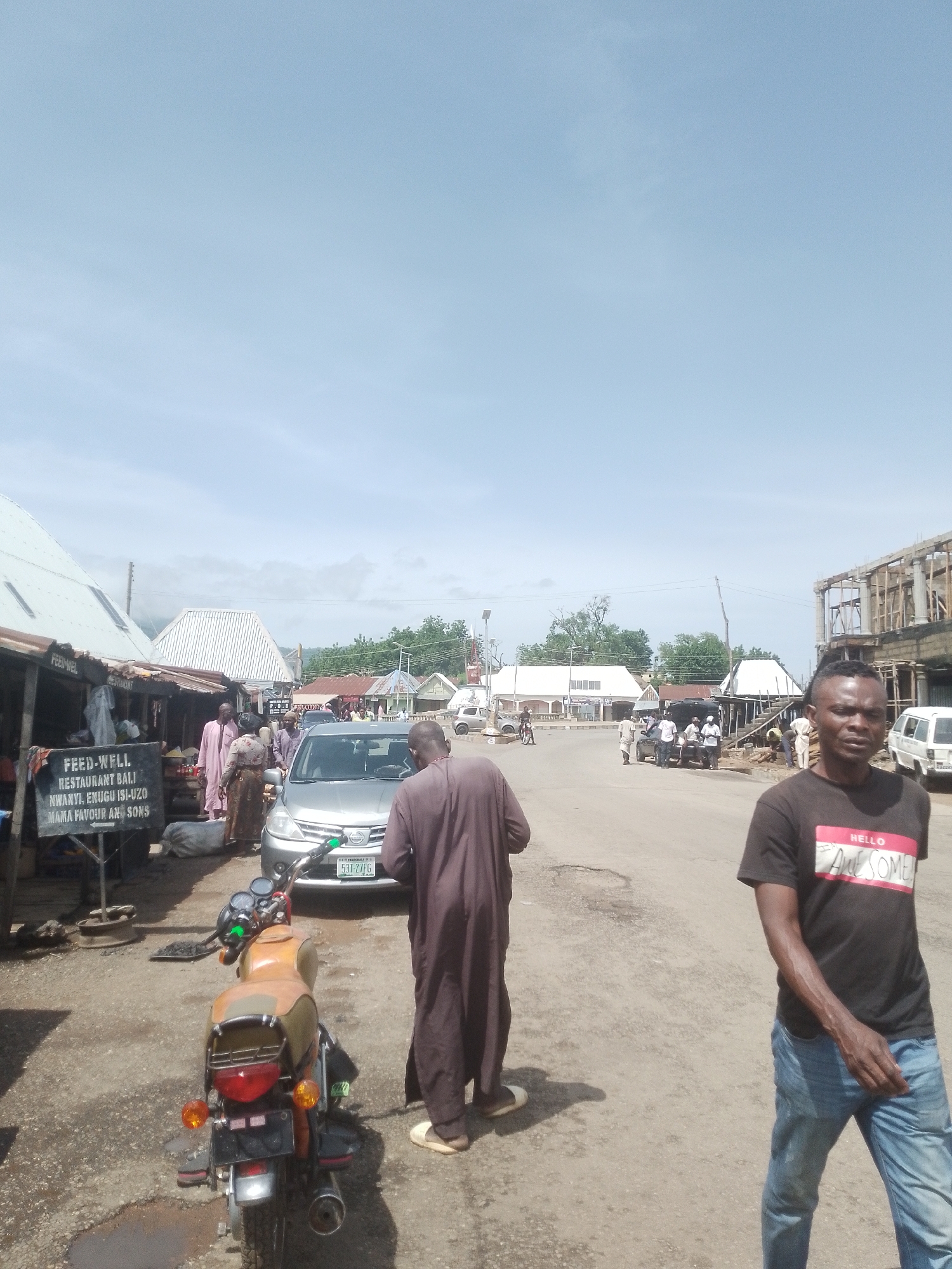This media file depicts a bustling street scene in Beli, Nigeria. The image features several individuals, including a man in the foreground wearing a black t-shirt with the text "HELLO AWESOME" and blue jeans, walking towards the viewer. To his left, another person in a brown robe walks away from the camera. In the background, a silver sedan is parked, and several other people are visible near shops and along the street. A motorcycle is parked in the lower left quadrant of the image.
The setting appears to be a market or commercial area, with various structures that resemble shops or stalls lining the street. One stall has a sign that reads "FEED-WELL RESTAURANT BALI NWANYI, ENUGU ISI-UZO MAMA FAVOUR AN SONS." The road is unpaved and appears somewhat worn. In the distance, more buildings and trees are visible, suggesting an urban or semi-urban environment. The sky is bright and clear, indicating it is daytime with fair weather. The overall impression is one of everyday activity and commerce. The presence of multiple people and vehicles suggests a busy period.
