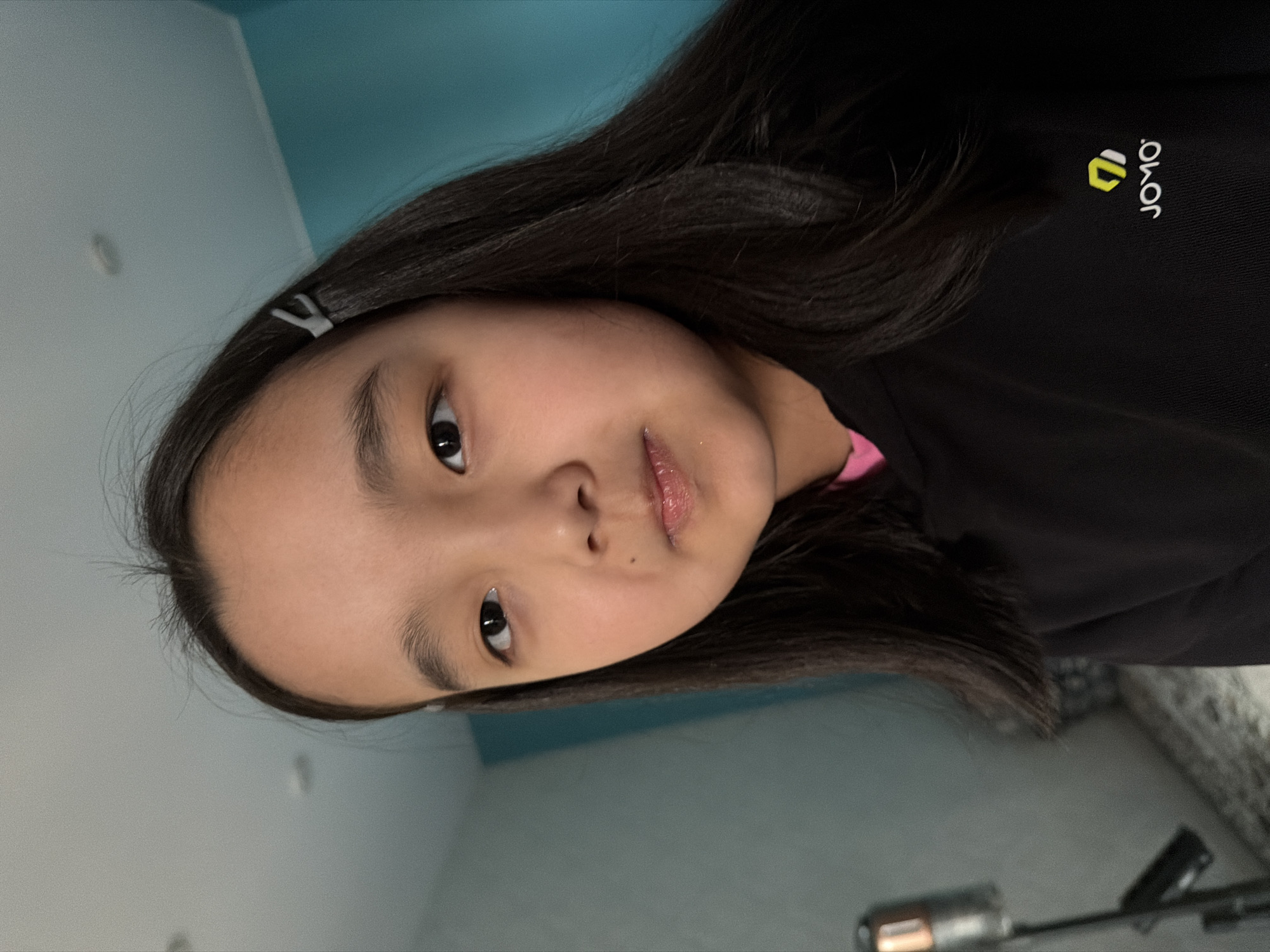 A close-up, head-and-shoulders selfie of a young Asian girl is shown. She has long, dark hair with a white clip on the right side. Her skin is fair, and she is looking directly at the camera with a neutral expression. She is wearing a black hoodie with a small yellow and white logo on the lower right side. The background is blurred, with a turquoise wall on the right and a grey wall on the left, suggesting an indoor setting. There is a faint reflection of a light source on her lips, indicating glossy or wet lips. The lighting is bright and direct, coming from above and slightly in front of her.