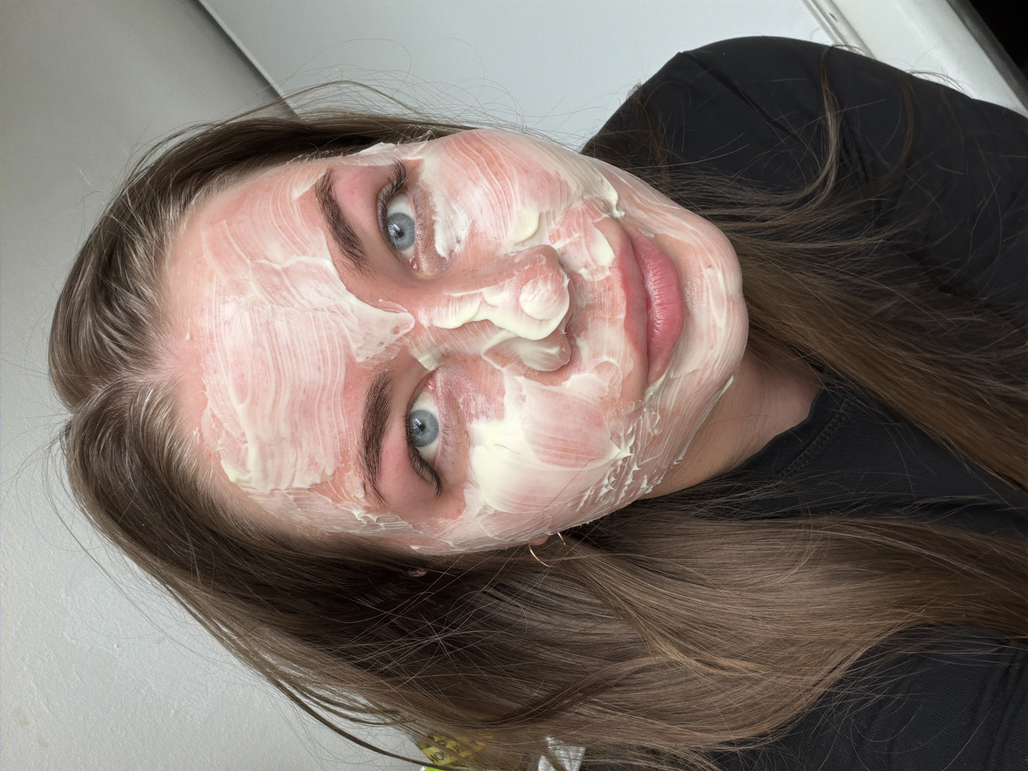 A young woman with light skin and blue eyes is looking directly at the camera. Her face is covered in a thick, white cream mask. She has brown hair that is parted to the side and falls around her face and shoulders. She is wearing a black shirt. The background is a plain white wall, possibly in a residential or domestic setting. There is no visible indication of the city of Chaikovsky, Russia.