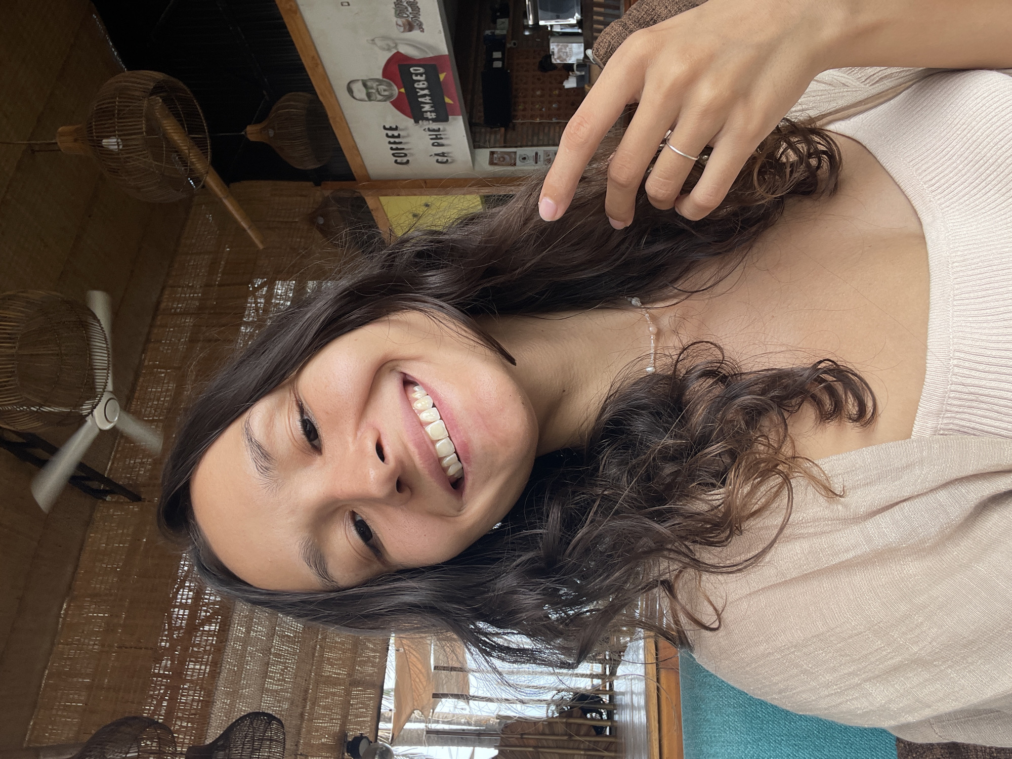 A young woman with long, dark, wavy hair smiles directly into the camera. She is wearing a light-colored, ribbed tank top and a sheer, beige cardigan. A delicate necklace with small beads is visible around her neck, and a thin silver ring adorns one finger. Her right hand is raised, with her fingers gently touching her hair. The background suggests an indoor cafe or restaurant setting with a rustic, tropical aesthetic. Woven bamboo lamps and a ceiling fan are visible overhead. A mural on the wall behind her depicts a cartoonish figure with the text "COFFEE CÀ PHÊ #MAXBEO." The overall scene appears casual and relaxed, potentially in Phu Thuy Ward, Vietnam.