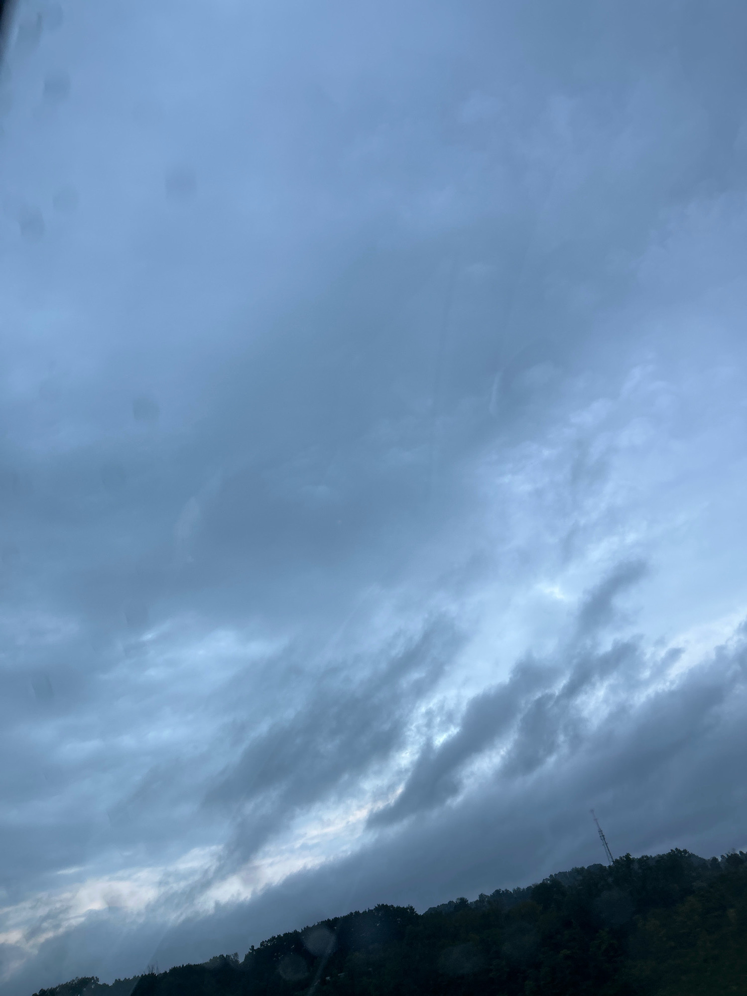 A low-angle, full shot captures a dark, overcast sky above a tree-lined hill. The sky is filled with heavy, gray clouds that appear to be moving, with some lighter patches suggesting breaks in the cloud cover. The clouds are textured, with streaks and wisps indicating movement and varying density. In the distance, silhouetted against the dim sky, is a dark green hillside covered in dense trees. Towards the right side of the frame, a tall, slender communication tower is visible, rising from the crest of the hill. The overall atmosphere is subdued and somewhat melancholic, suggesting either impending rain or the aftermath of a storm. The image appears to have been taken from a moving vehicle, as evidenced by some water spots or smudges on the foreground, possibly on a window. The location is identified as Collier Township, United States of America.