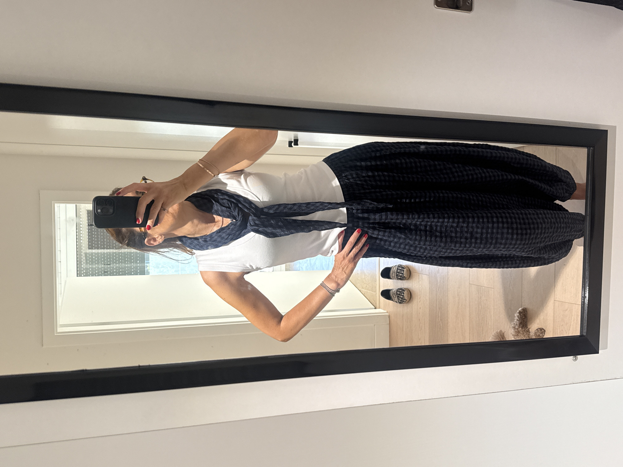 A woman with brown hair, wearing a white tank top, a dark blue scarf tied around her neck, and dark checkered pants, takes a selfie in a full-length mirror. She is also wearing a thin bracelet on her left wrist and has red nail polish on her fingernails. Her pose suggests confidence, with one hand on her hip. In the reflection of the mirror, a pair of patterned slip-on shoes are visible on the floor, along with a small, light brown dog. The environment appears to be an indoor setting, possibly a bedroom or dressing area, with light-colored wooden flooring. The overall impression is a casual and stylish moment captured in a personal space. The image was taken in Loures, Portugal.