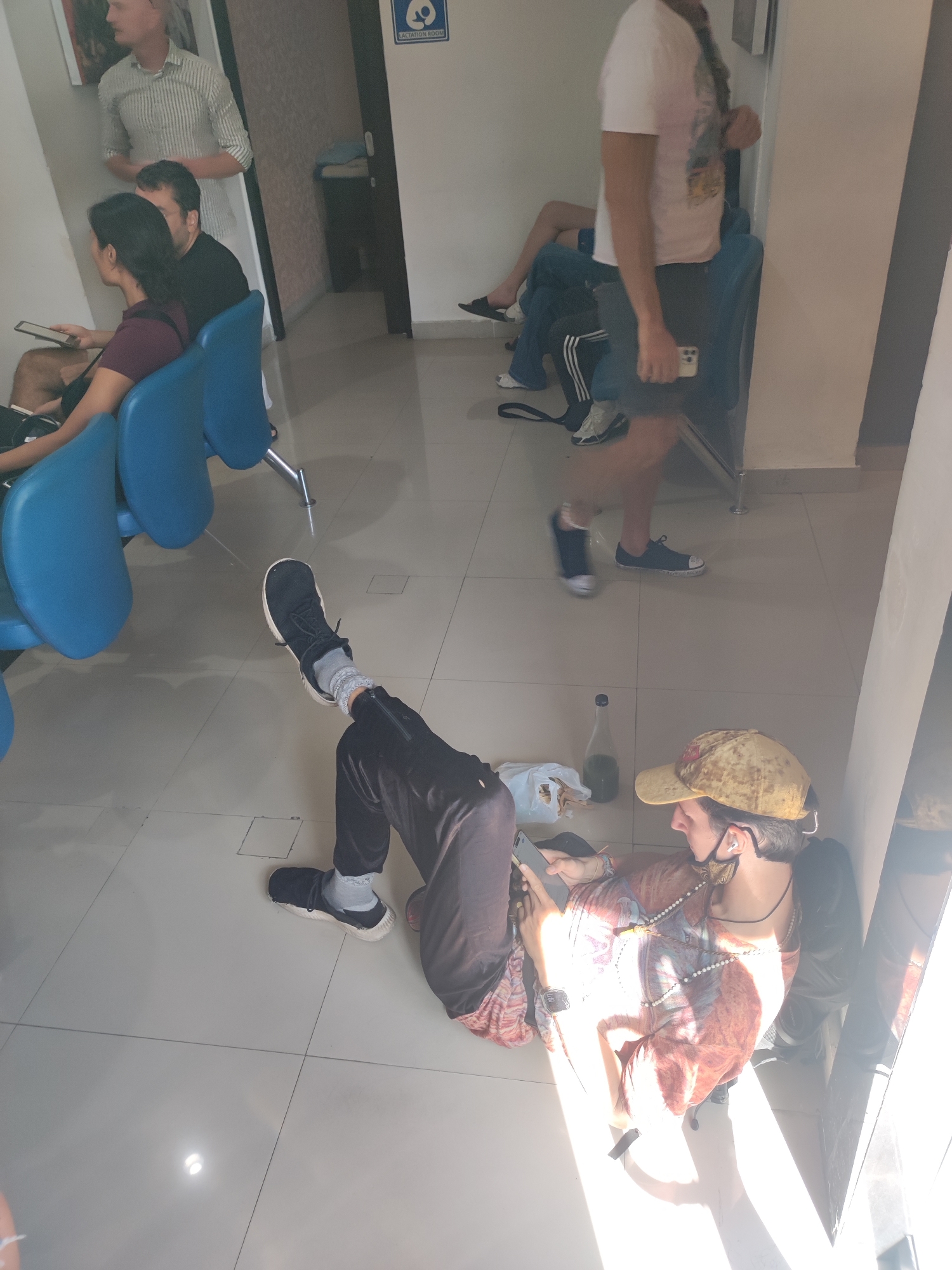 The photo shows a waiting area in a building in Denpasar, Indonesia. The floor is tiled and reflective.
Several people are present. Three people are sitting on blue, connected chairs. A man in a striped shirt stands behind them. Another man wearing a white shirt and shorts walks past. On the floor, a person wearing a patterned shirt, black pants, and a yellow cap lies against a wall, looking at a phone. Next to this person is a plastic bag, a bottle, and a dark object.